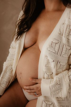 Intimate maternity photo showing pregnant belly with white knit cardigan against warm brown background.