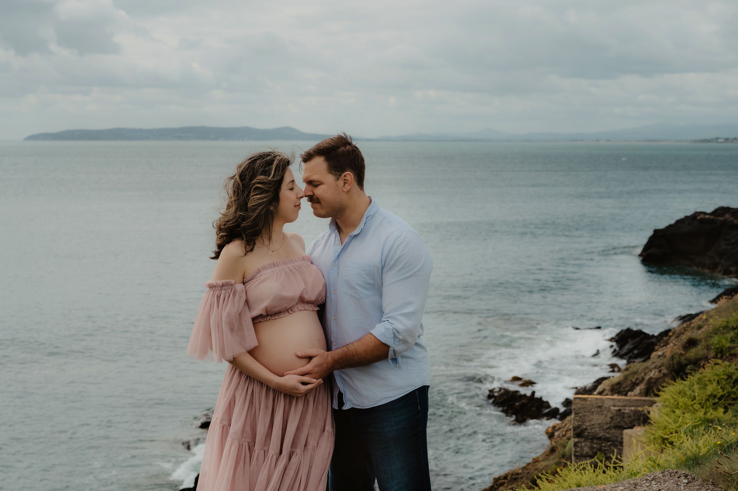 Expecting couple shares tender moment on cliff overlooking ocean vista.