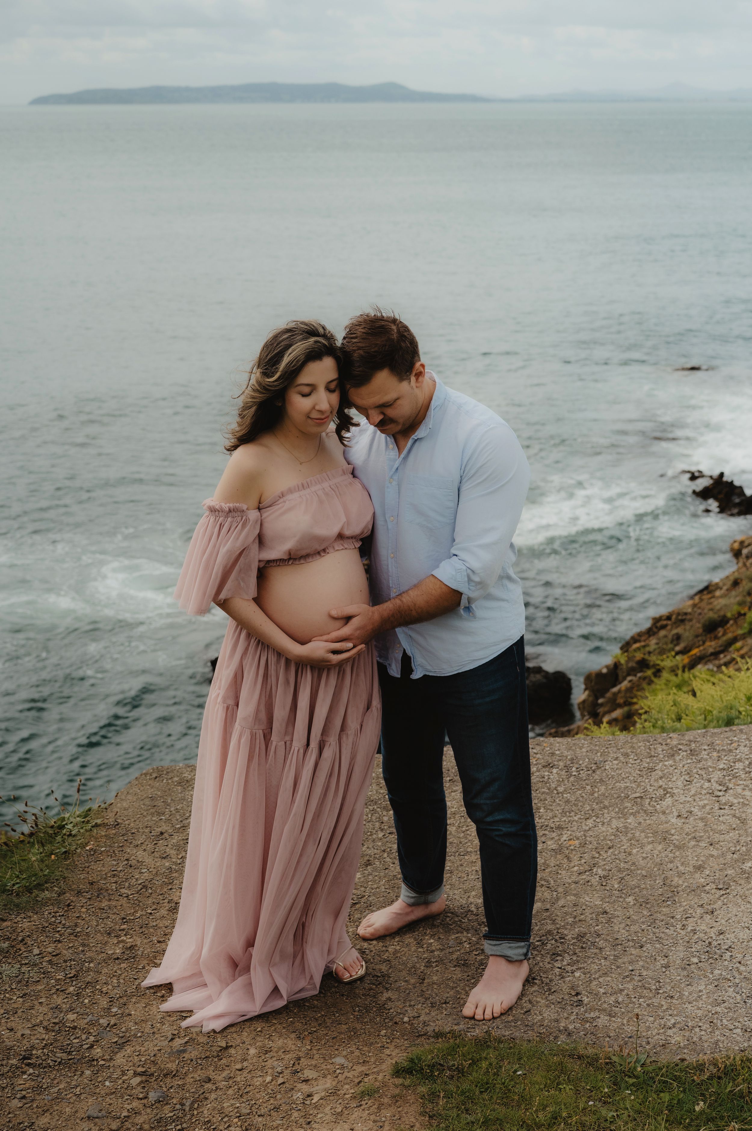 Barefoot couple embraces during sunset maternity shoot on coastal cliff.