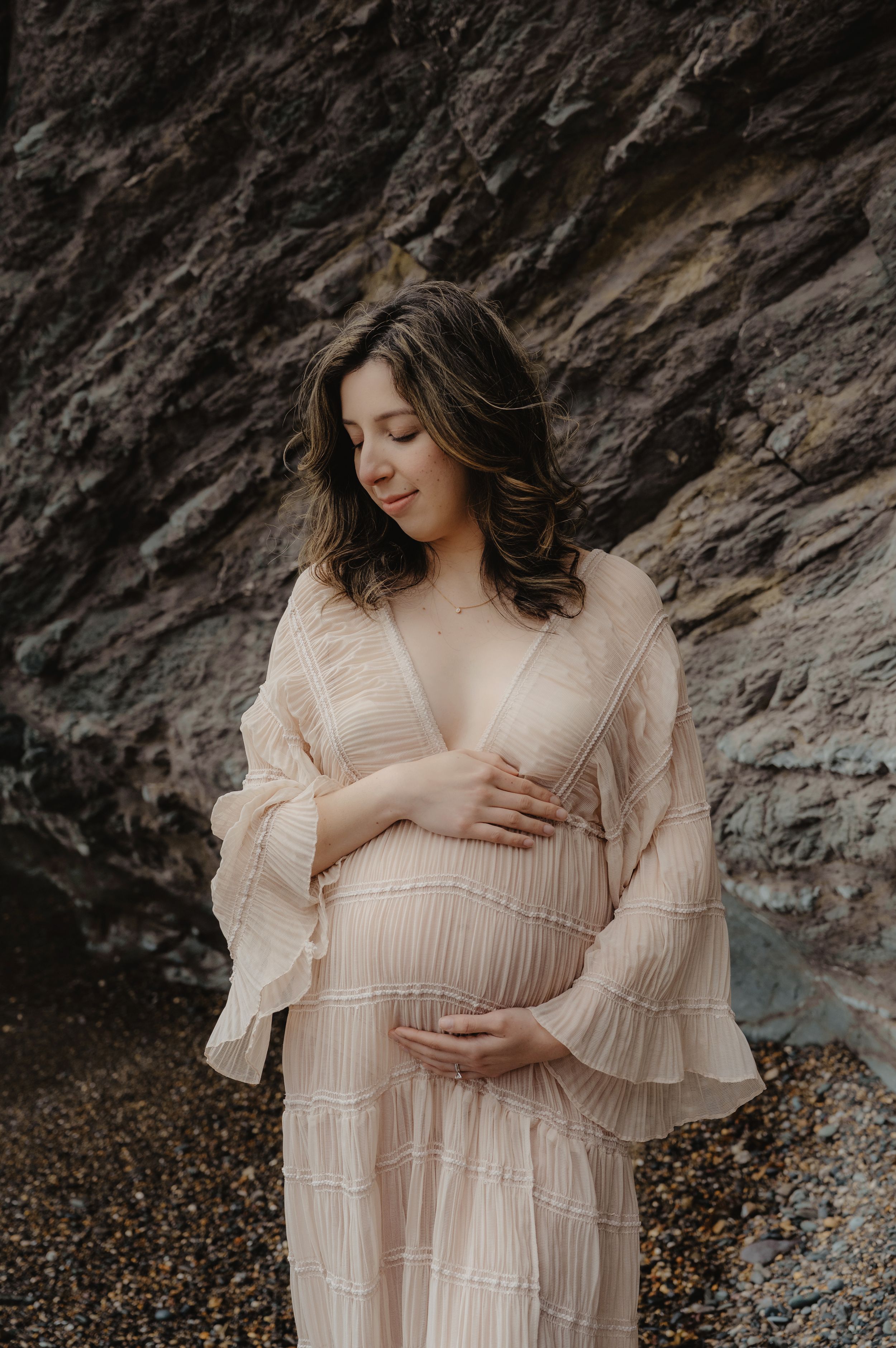 Expectant mother in delicate beige dress poses against rocky beach backdrop.
