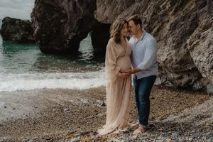 Romantic maternity photo of couple standing under coastal rock formation.