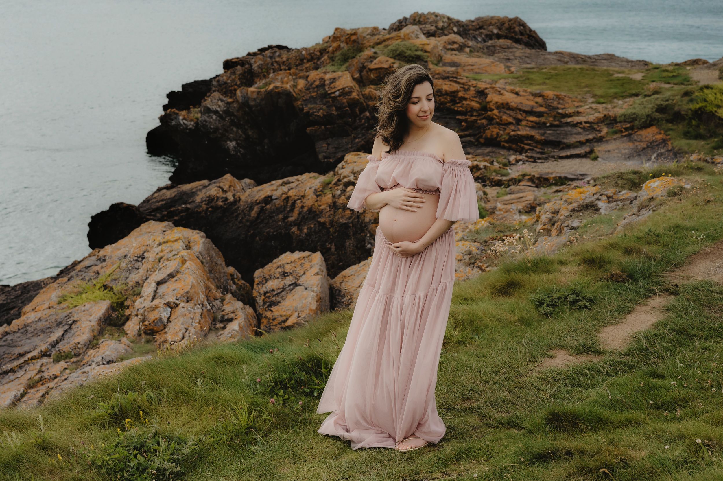 Mother-to-be in pink flowing dress poses on dramatic coastal cliffs.