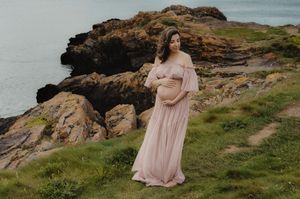 Mother-to-be in pink flowing dress poses on dramatic coastal cliffs.