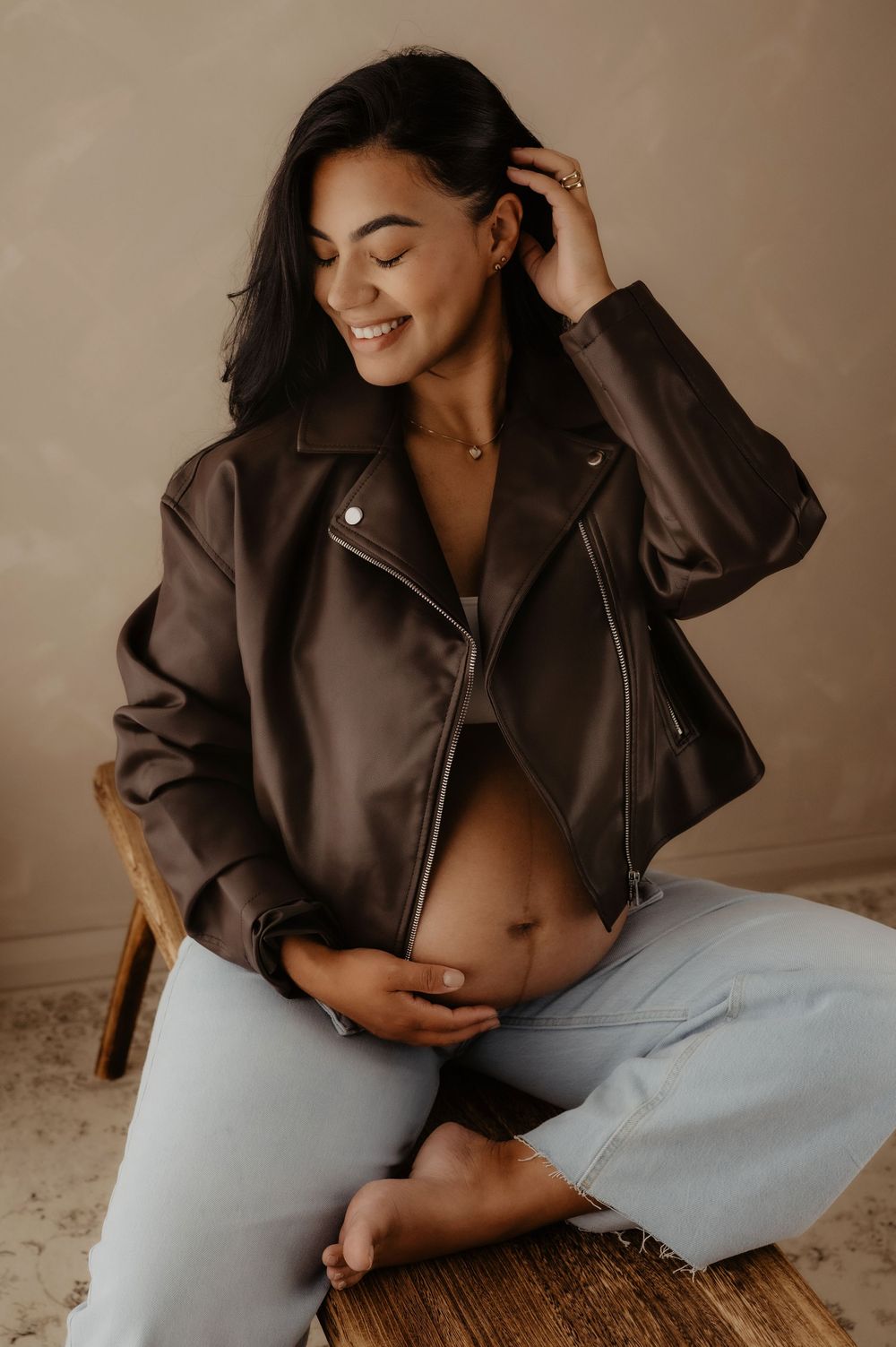 Casual maternity photo with woman in brown leather jacket touching her hair while seated on wooden bench.
