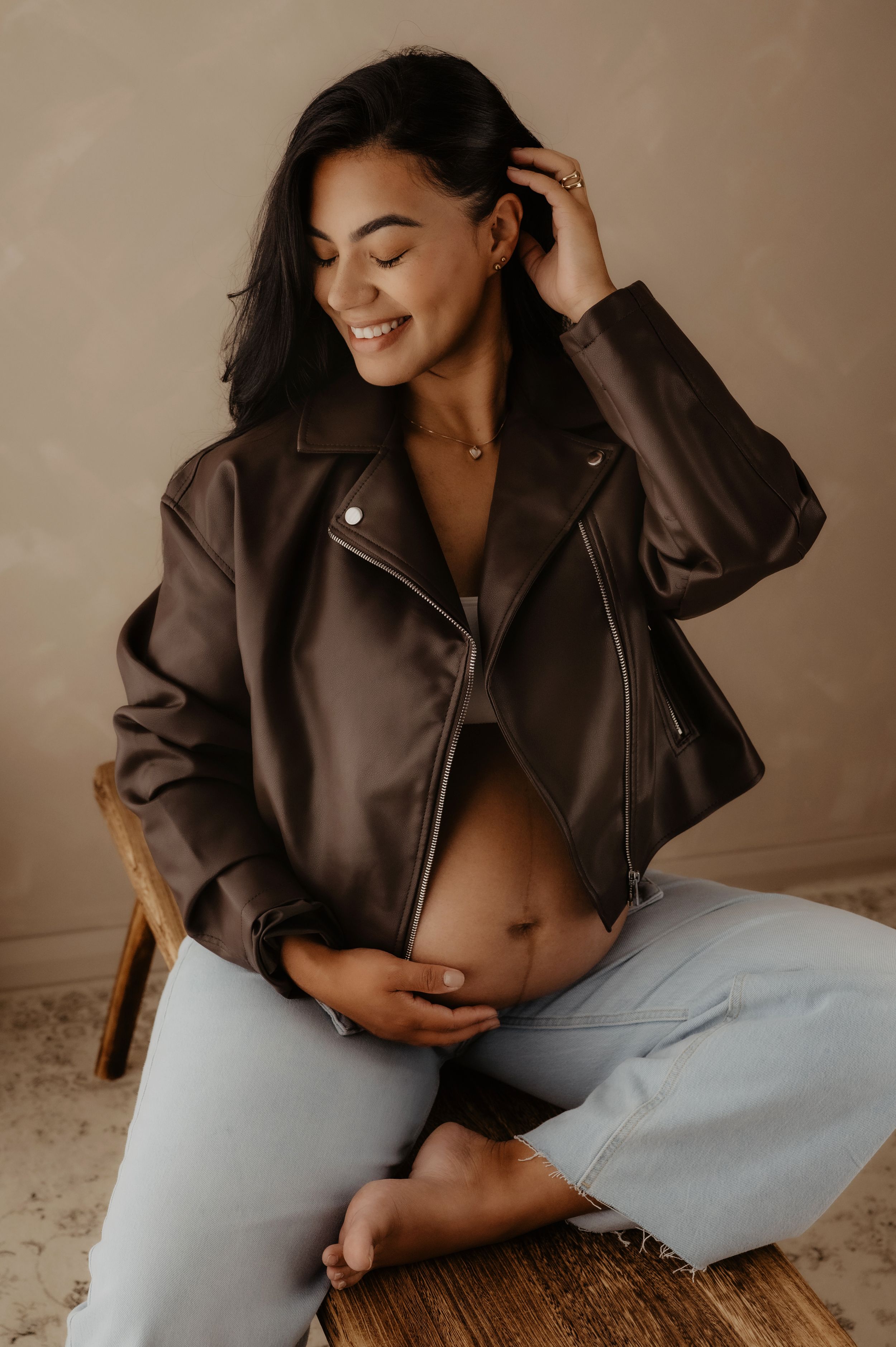 Casual maternity photo with woman in brown leather jacket touching her hair while seated on wooden bench.