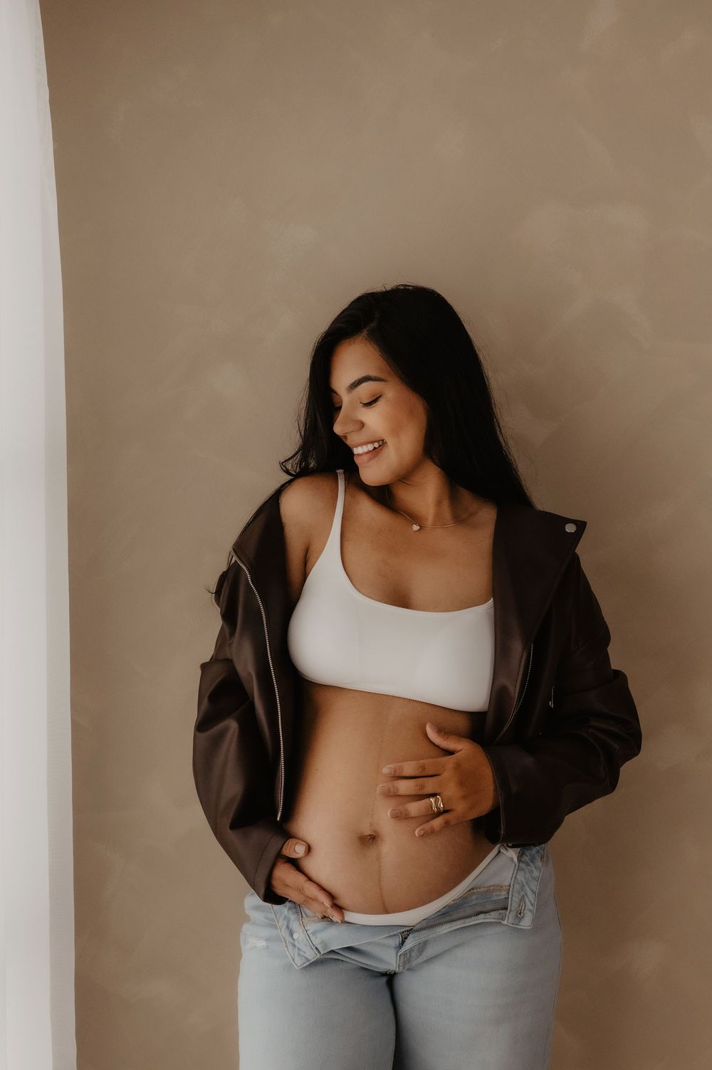 A maternity photo shows a woman in a white crop top and leather jacket posing against a beige wall.