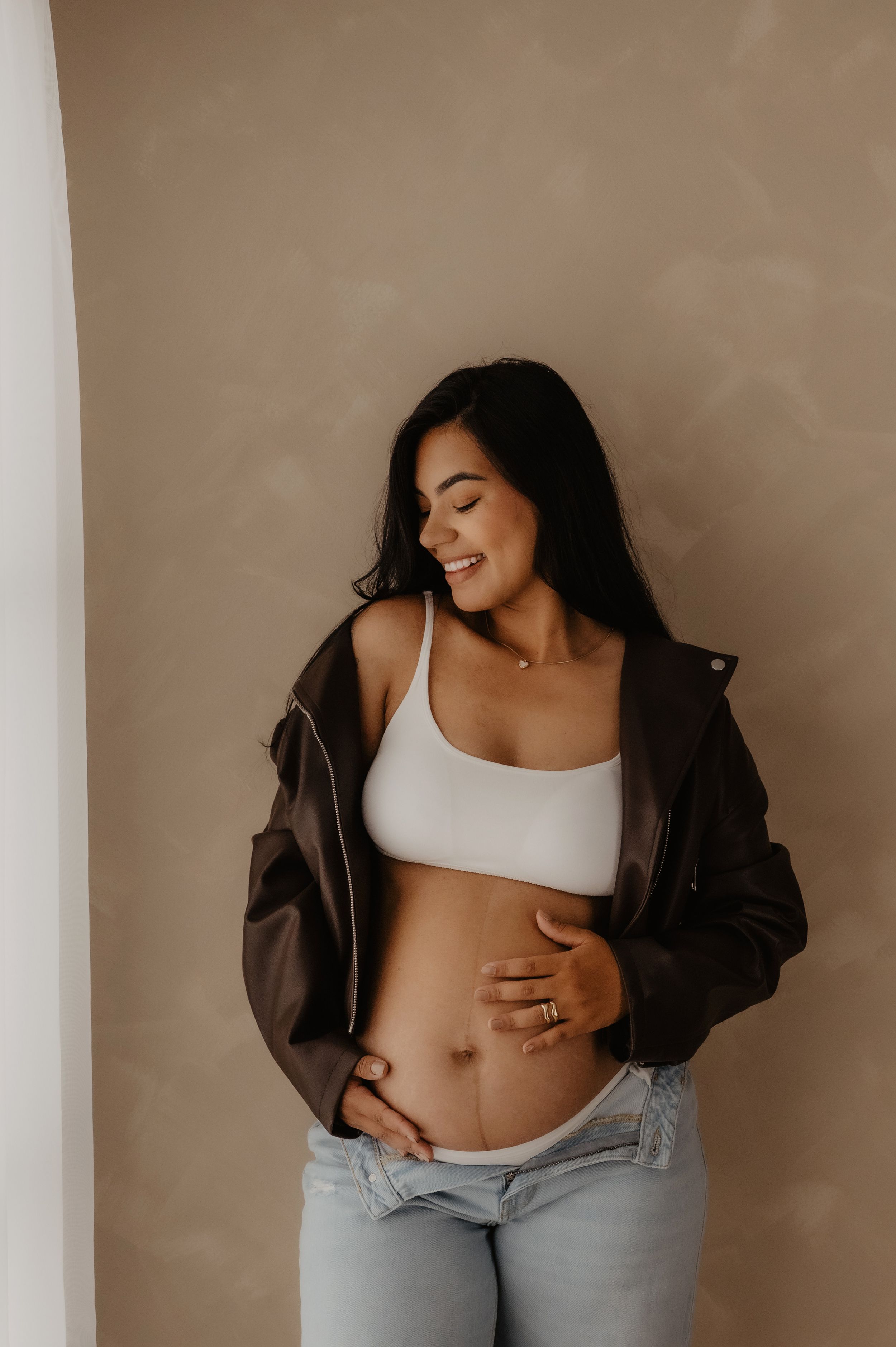 A maternity photo shows a woman in a white crop top and leather jacket posing against a beige wall.
