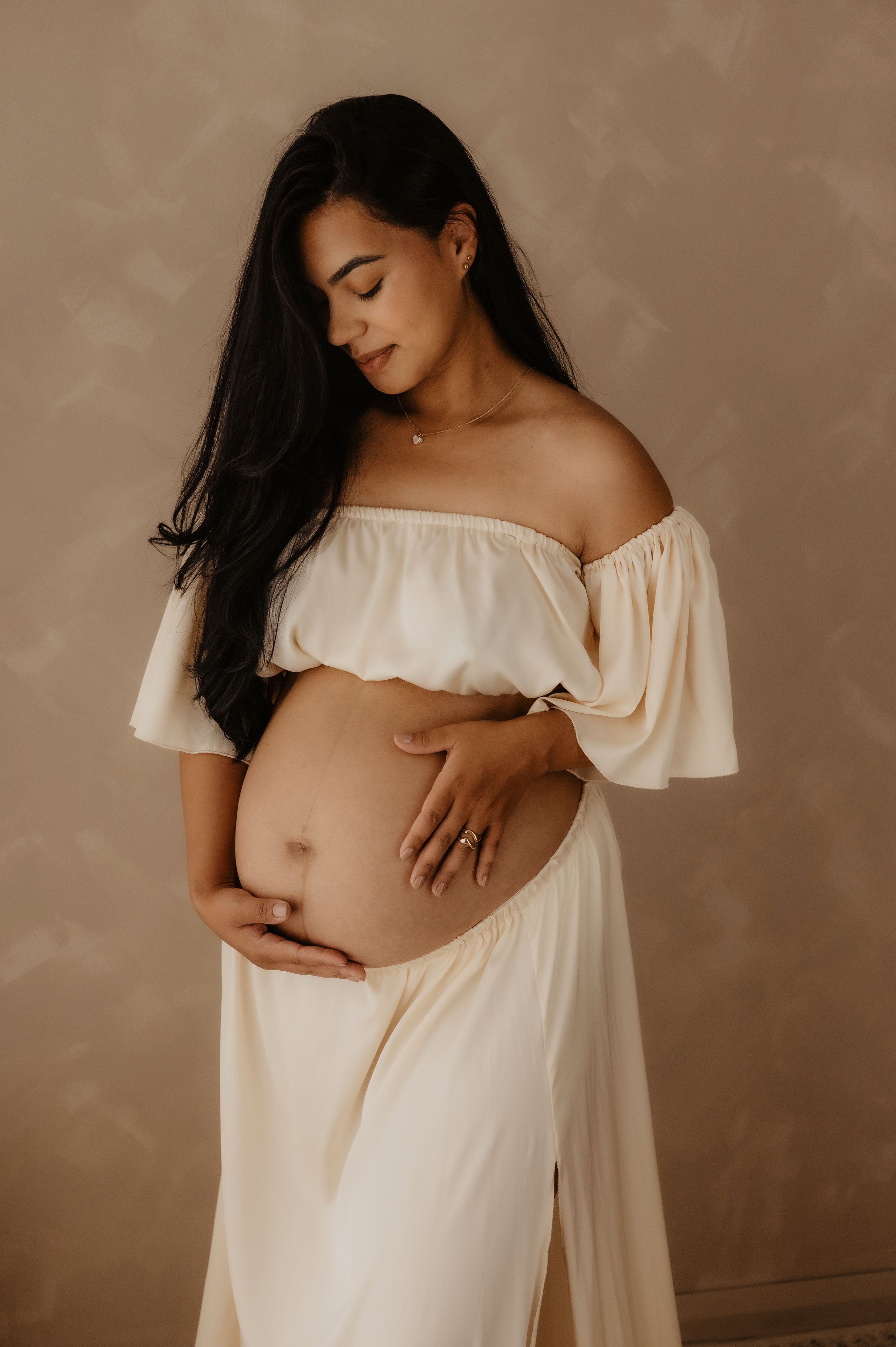 Expectant mother in flowing off-shoulder white dress poses tenderly with hands on belly in warm studio portrait.