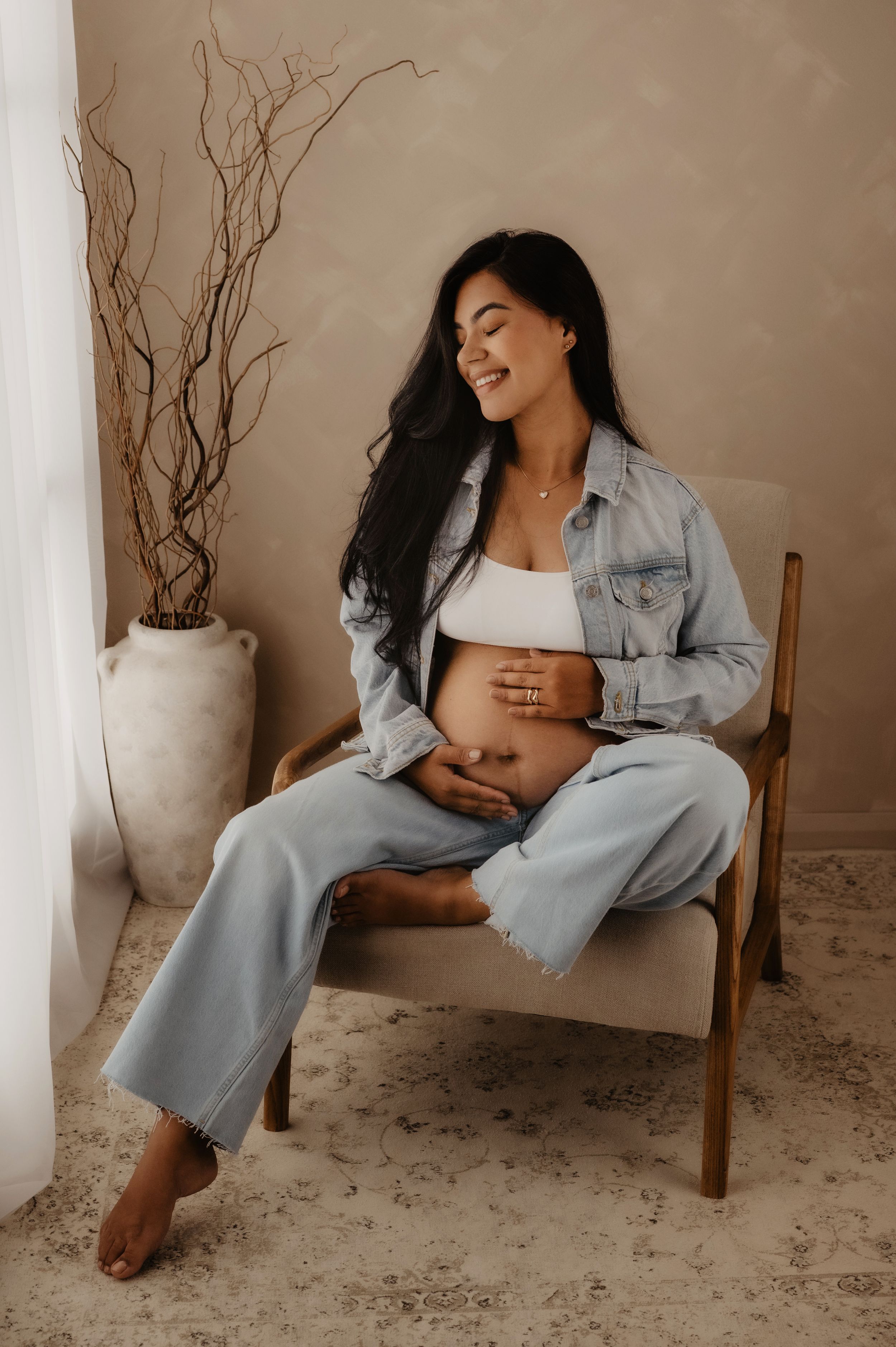 Expectant mother in denim jacket and jeans sits barefoot on chair while touching her belly with a smile.