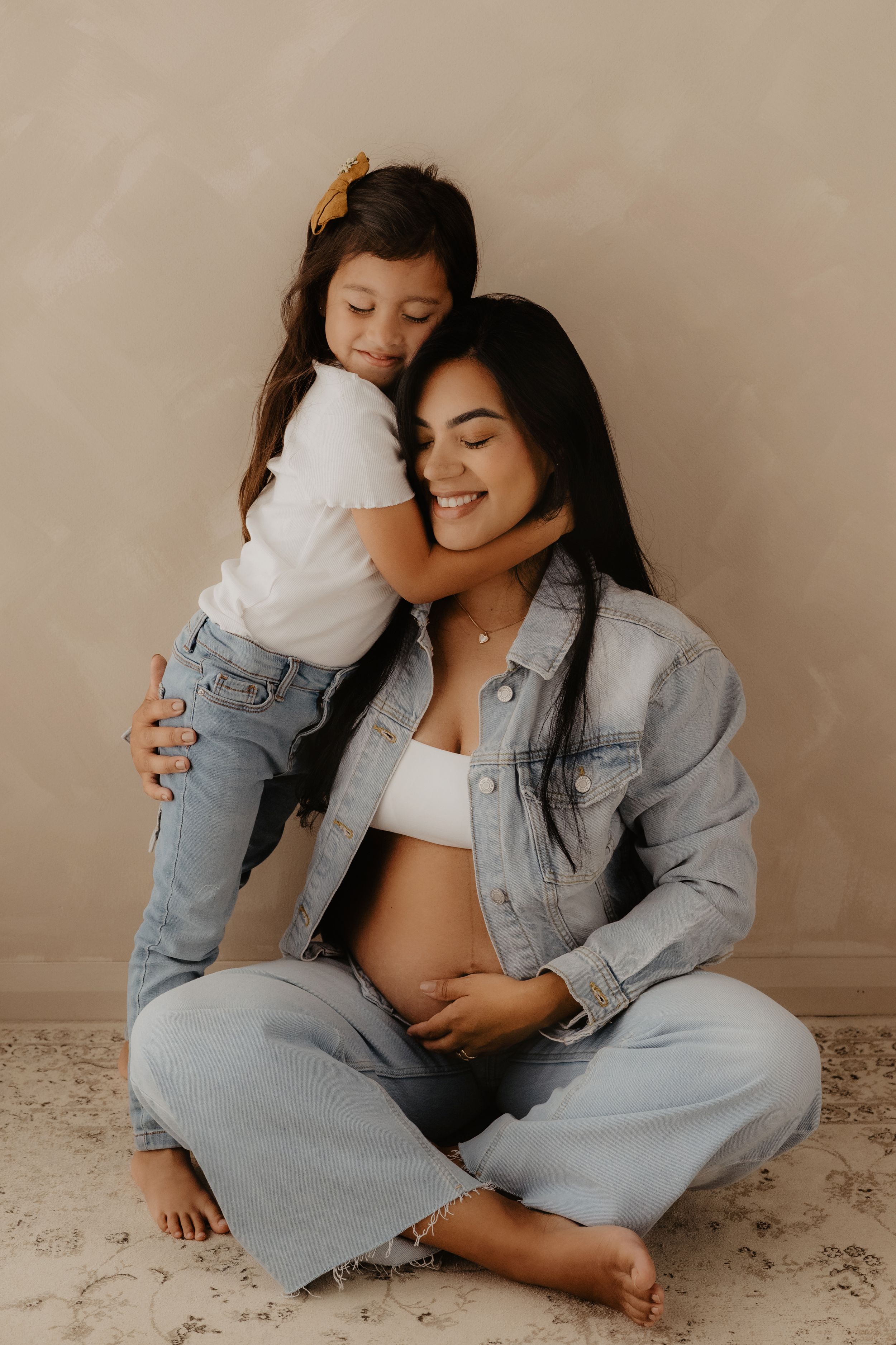 An intimate maternity photoshoot showing a tender embrace between mother and child against a neutral backdrop.