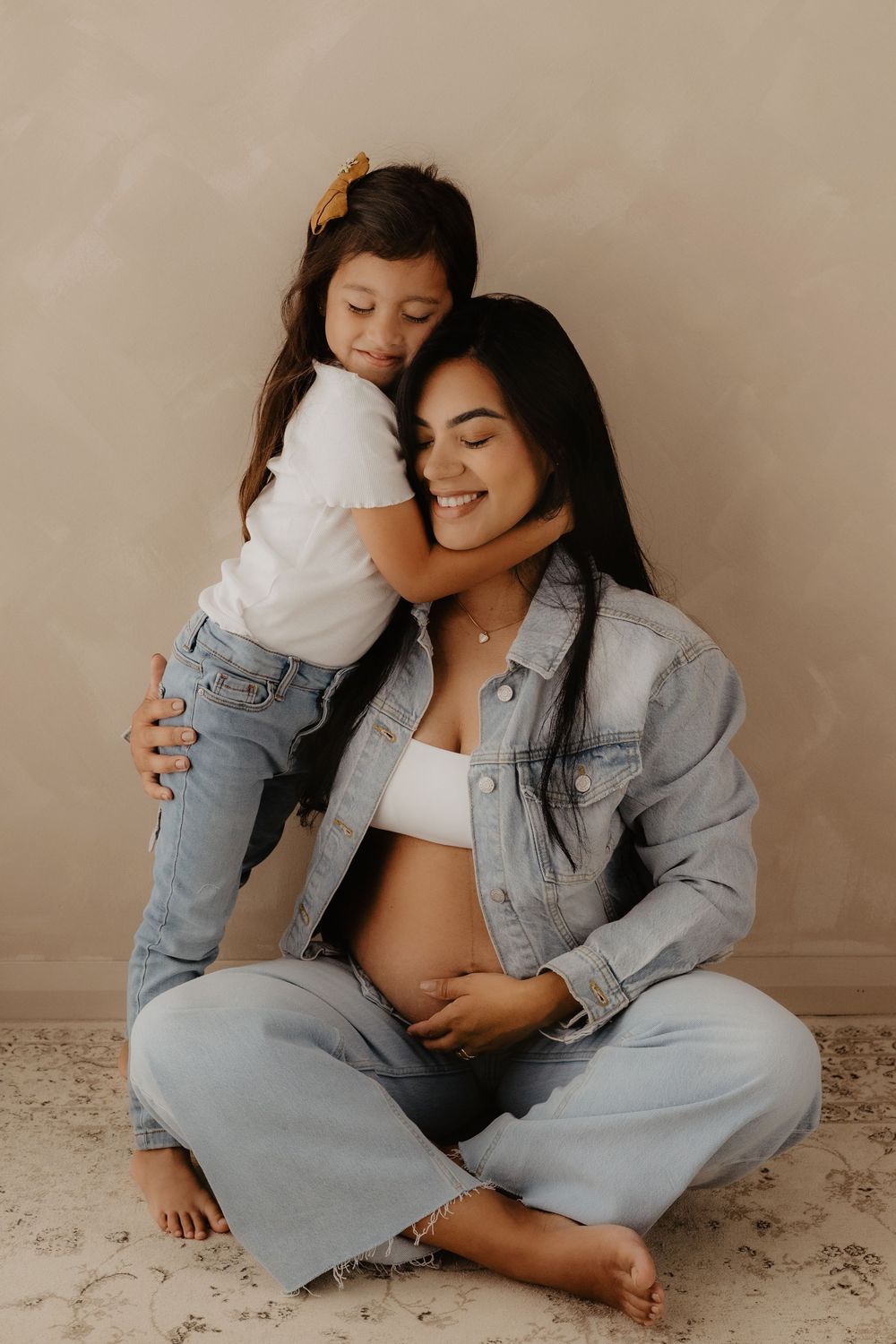 An intimate maternity photoshoot showing a tender embrace between mother and child against a neutral backdrop.