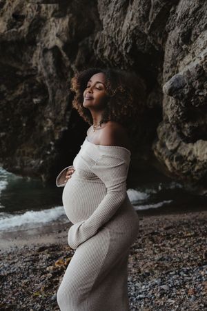 Expecting mother in cream knit dress poses against rocky coastal backdrop at sunset.