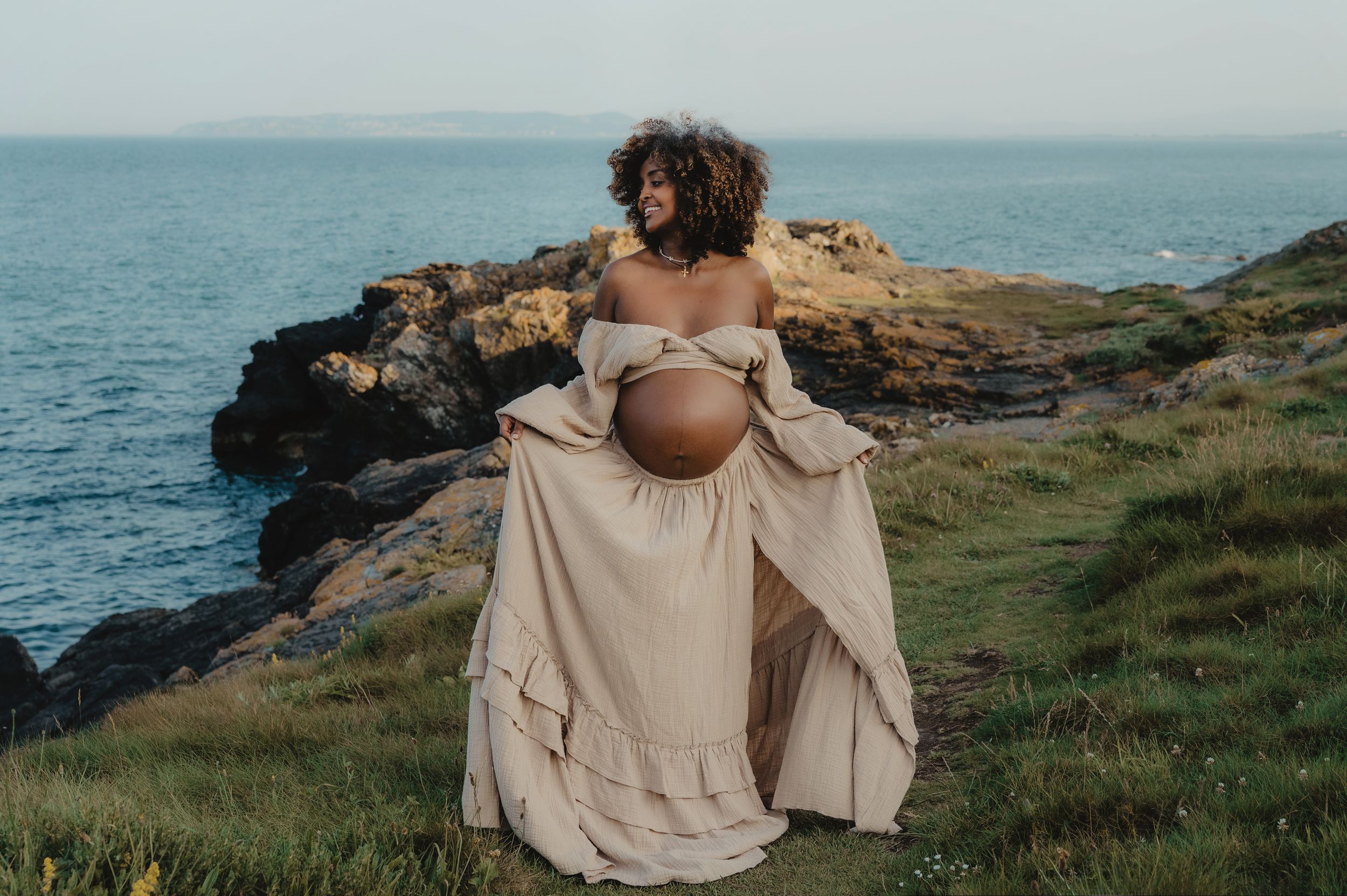 A beige dress billows in the wind during a clifftop maternity photograph.