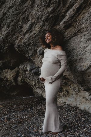 A maternity portrait against rocky textures wearing an off-shoulder cream dress.
