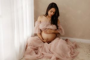 Serene maternity portrait of a woman in pink tulle dress sitting by window light.