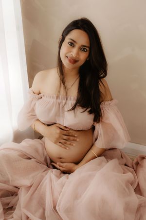 Maternity portrait of a woman in a pink dress cradling her baby bump against neutral background.