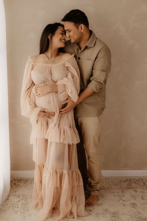 A romantic maternity portrait of a couple in neutral tones against a light wall.
