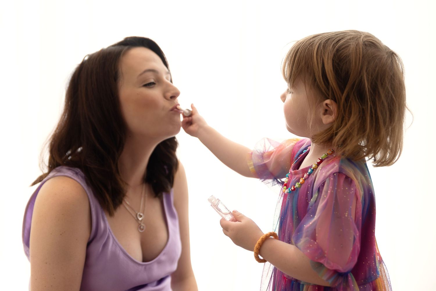 A toddler in pink clothing reaches out to touch their caregiver's face in a tender moment against white background.