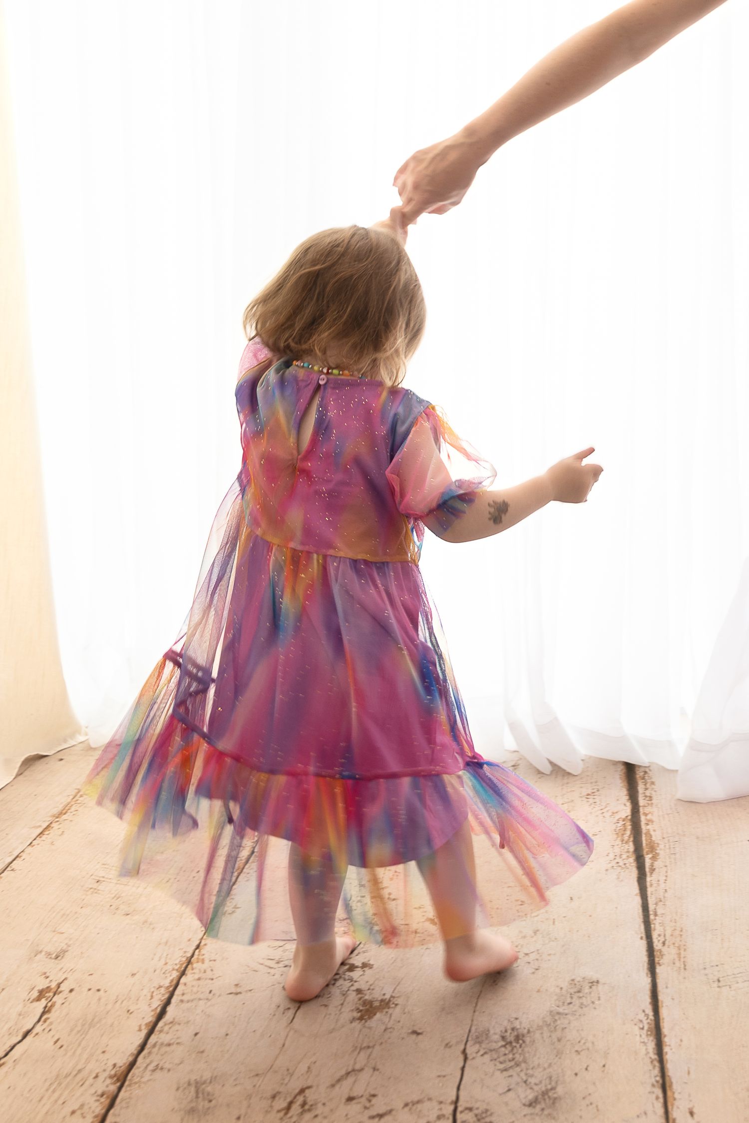 A child in a colorful purple tie-dye dress twirls and dances on a rustic wooden floor while a hand reaches down from above.