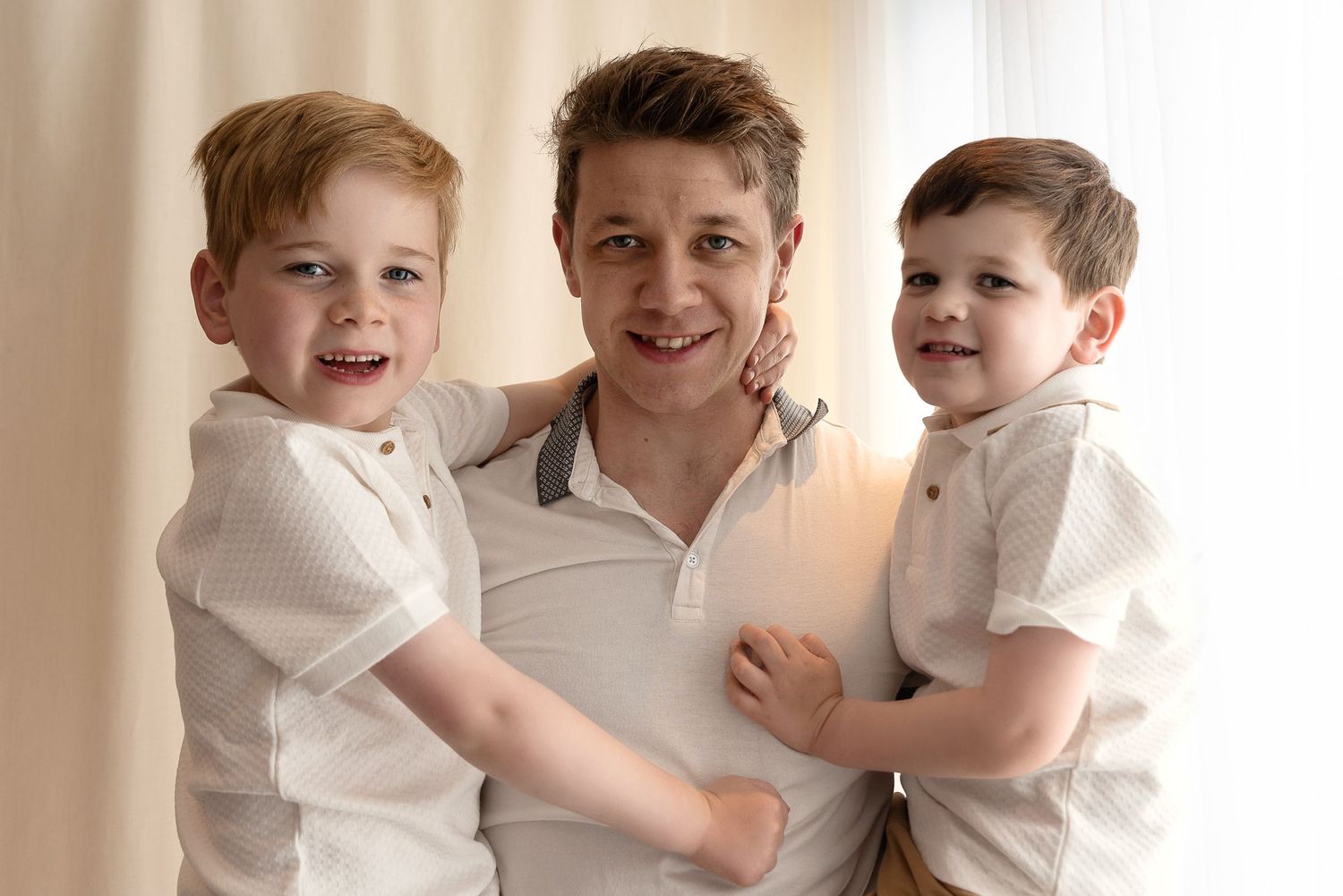 Three people wearing white polo shirts smile and embrace while posing together against a light colored background.