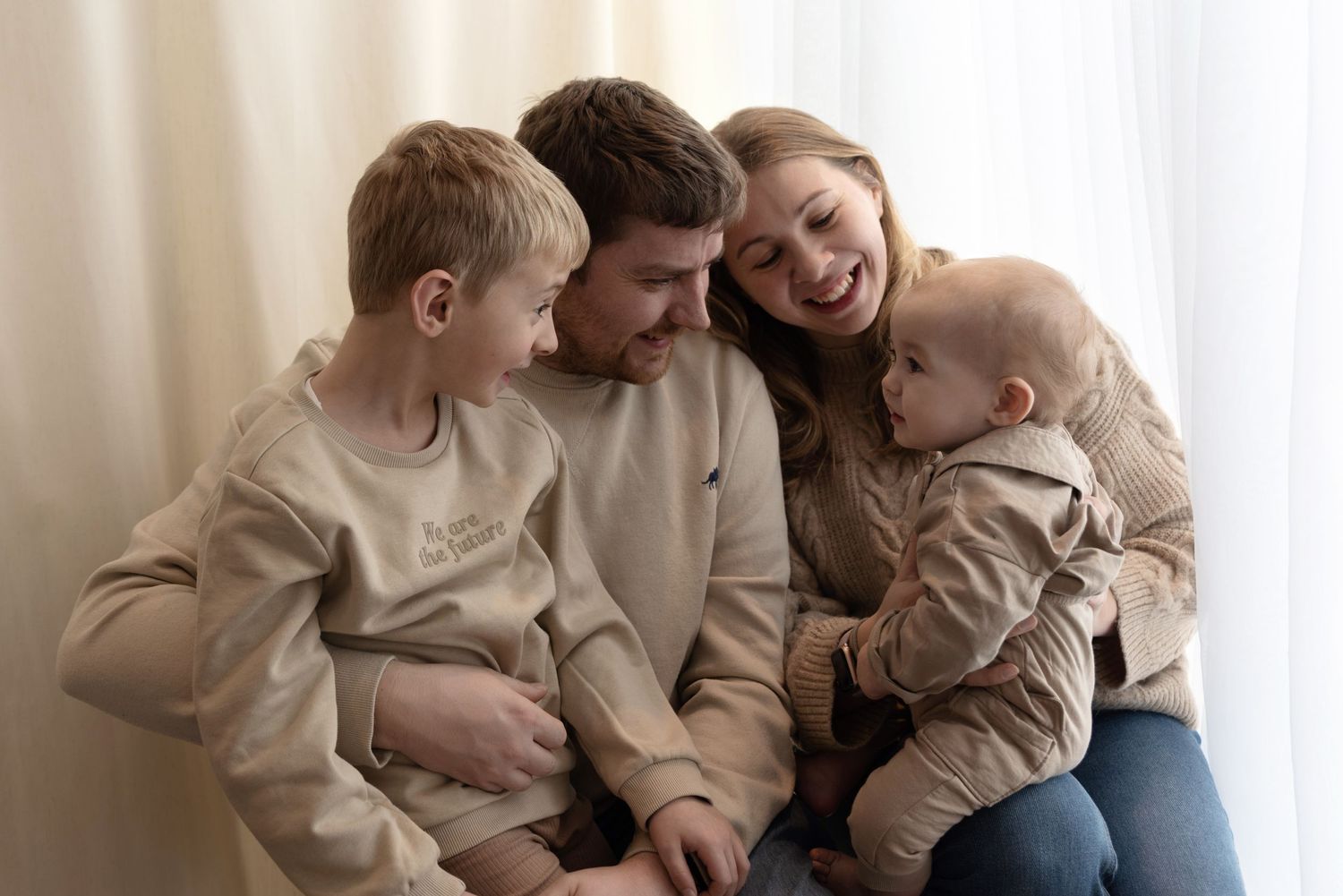 Happy family sitting together on couch at home wearing casual cream colored clothing and sharing joyful moment.