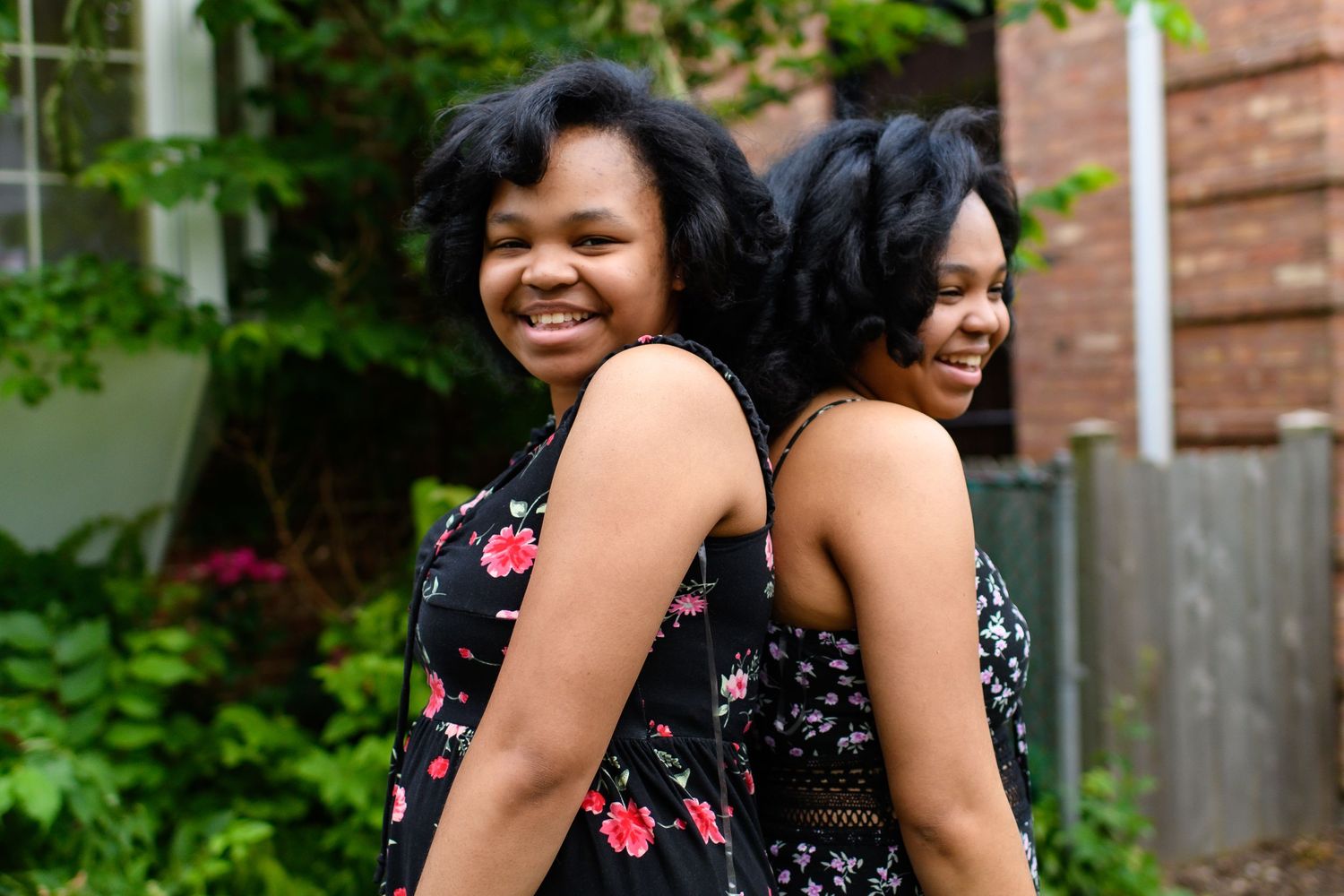 Two women in floral dresses stand back to back outdoors while smiling and laughing in a garden setting.
