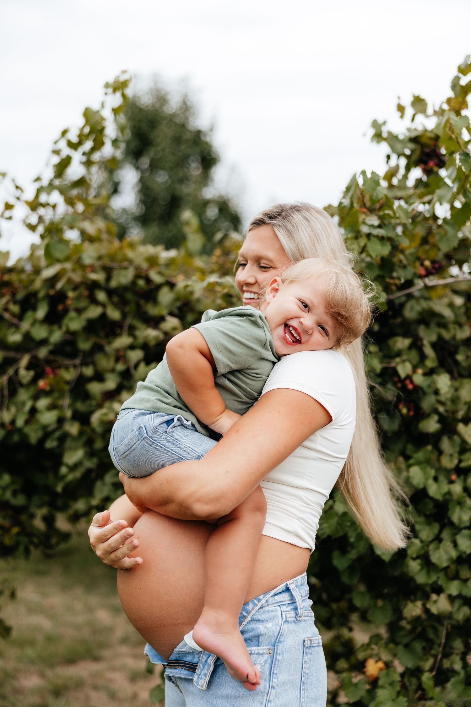 A smiling blonde mother holds her laughing young child outdoors among green berry bushes.