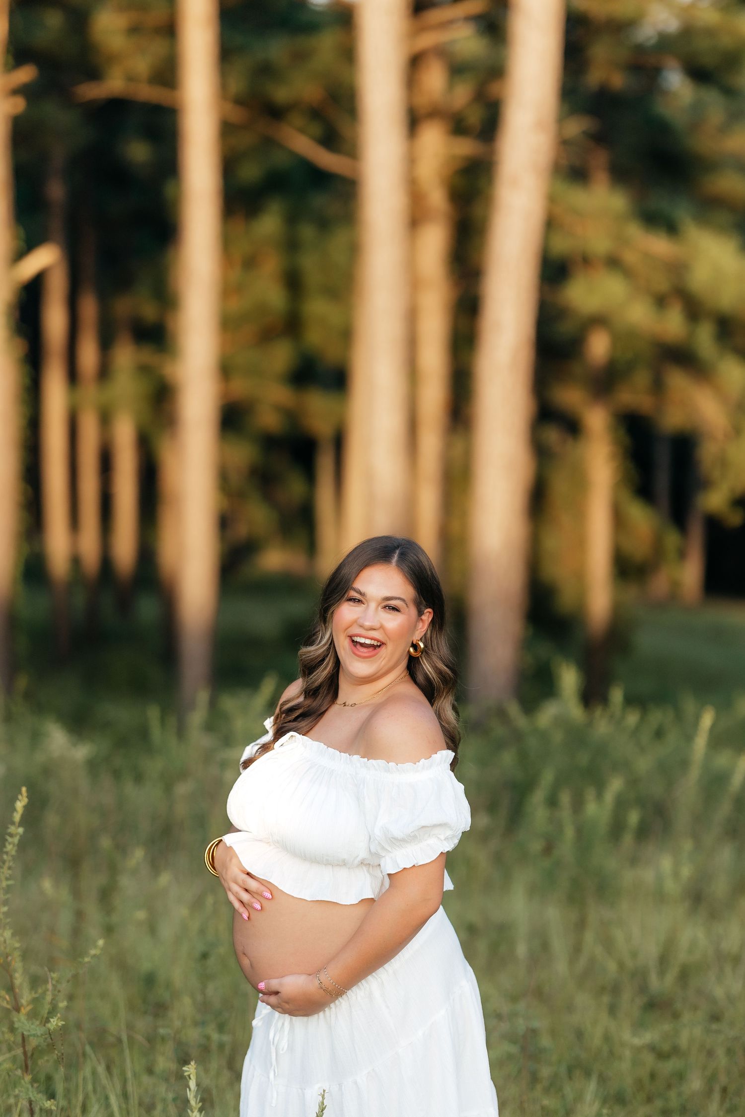 Woman in white off-shoulder dress laughs joyfully in a sunlit forest setting with tall pine trees in background.