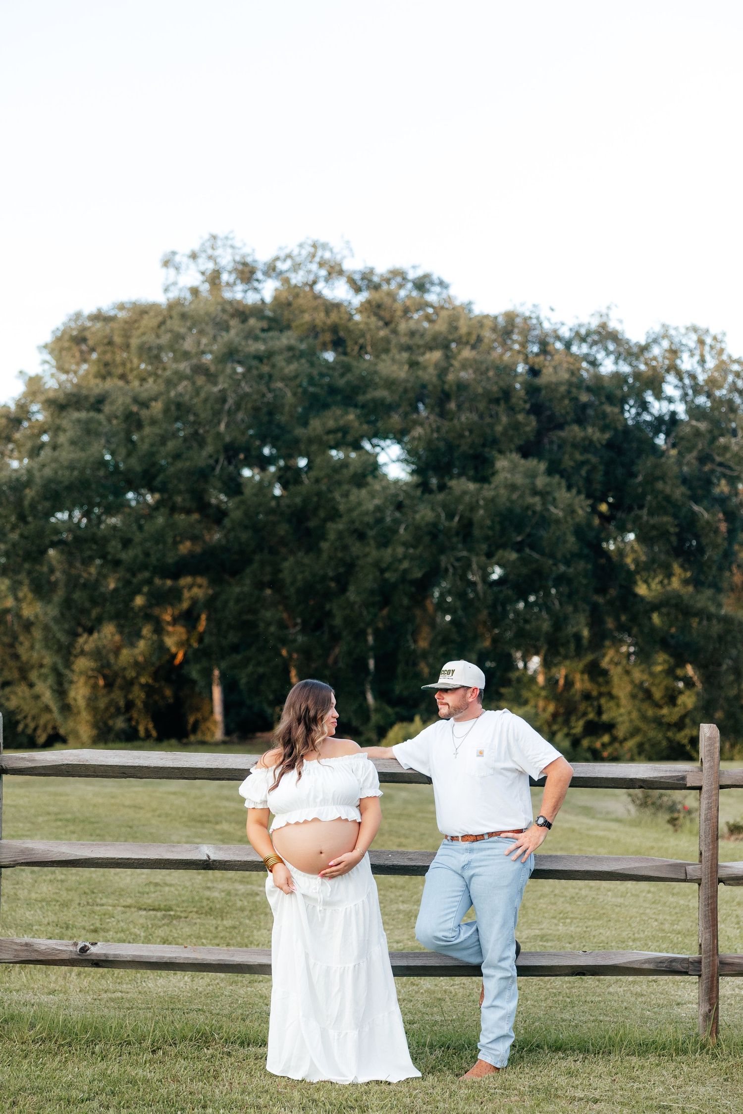 Couple in white and blue outfits walking along a wooden fence in a grassy field with trees in the background.