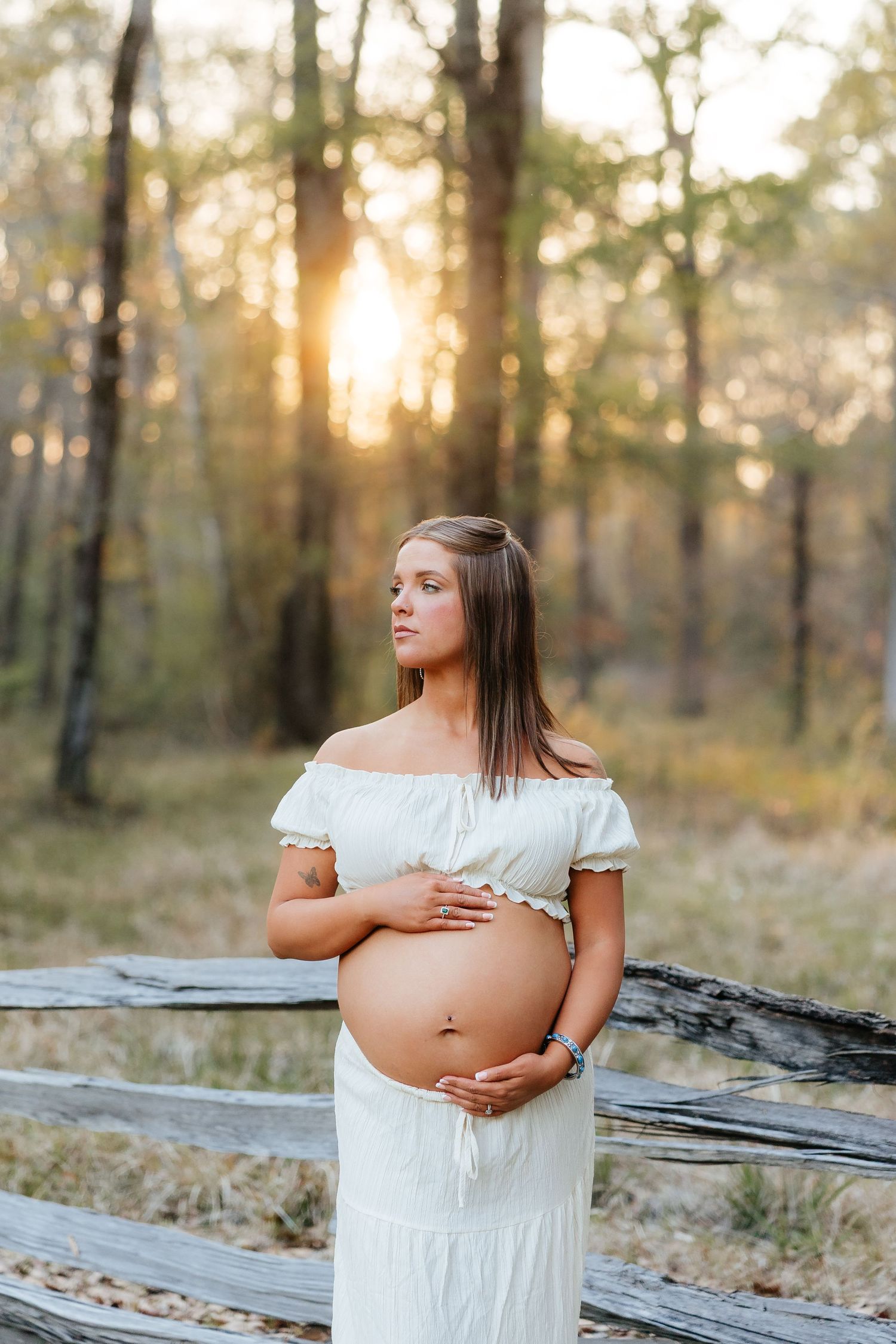 Pregnant woman in white off-shoulder dress holding baby bump at golden hour near rustic fence in forest setting.