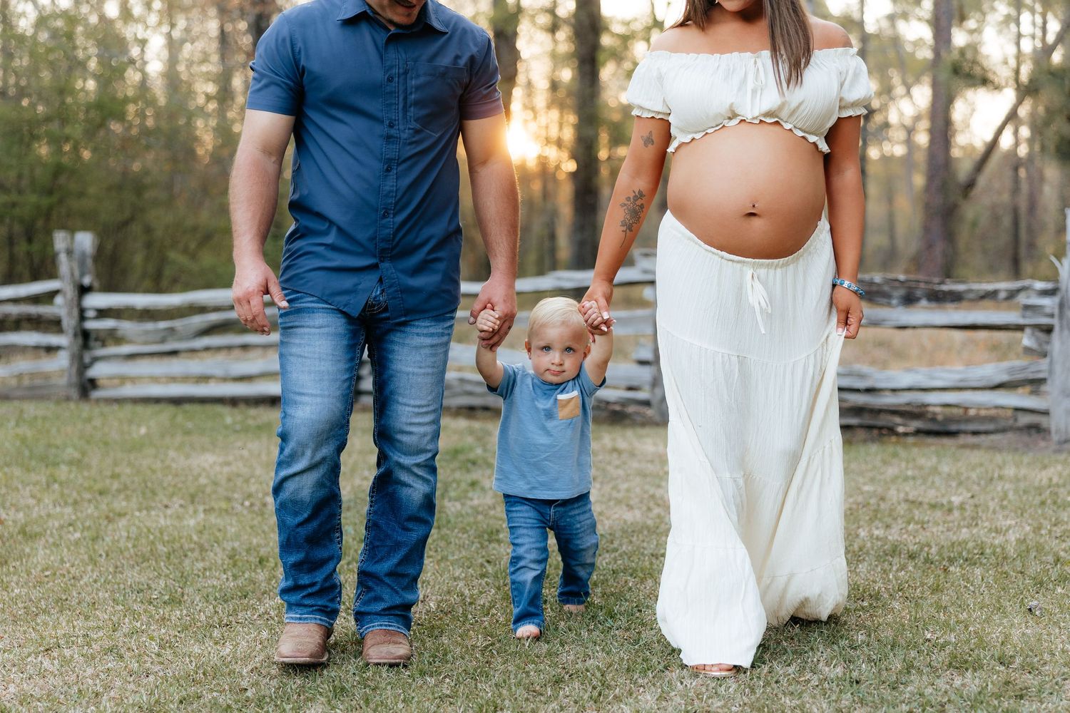 Pregnant woman and man hold toddler's hands during golden hour maternity photo session outdoors.