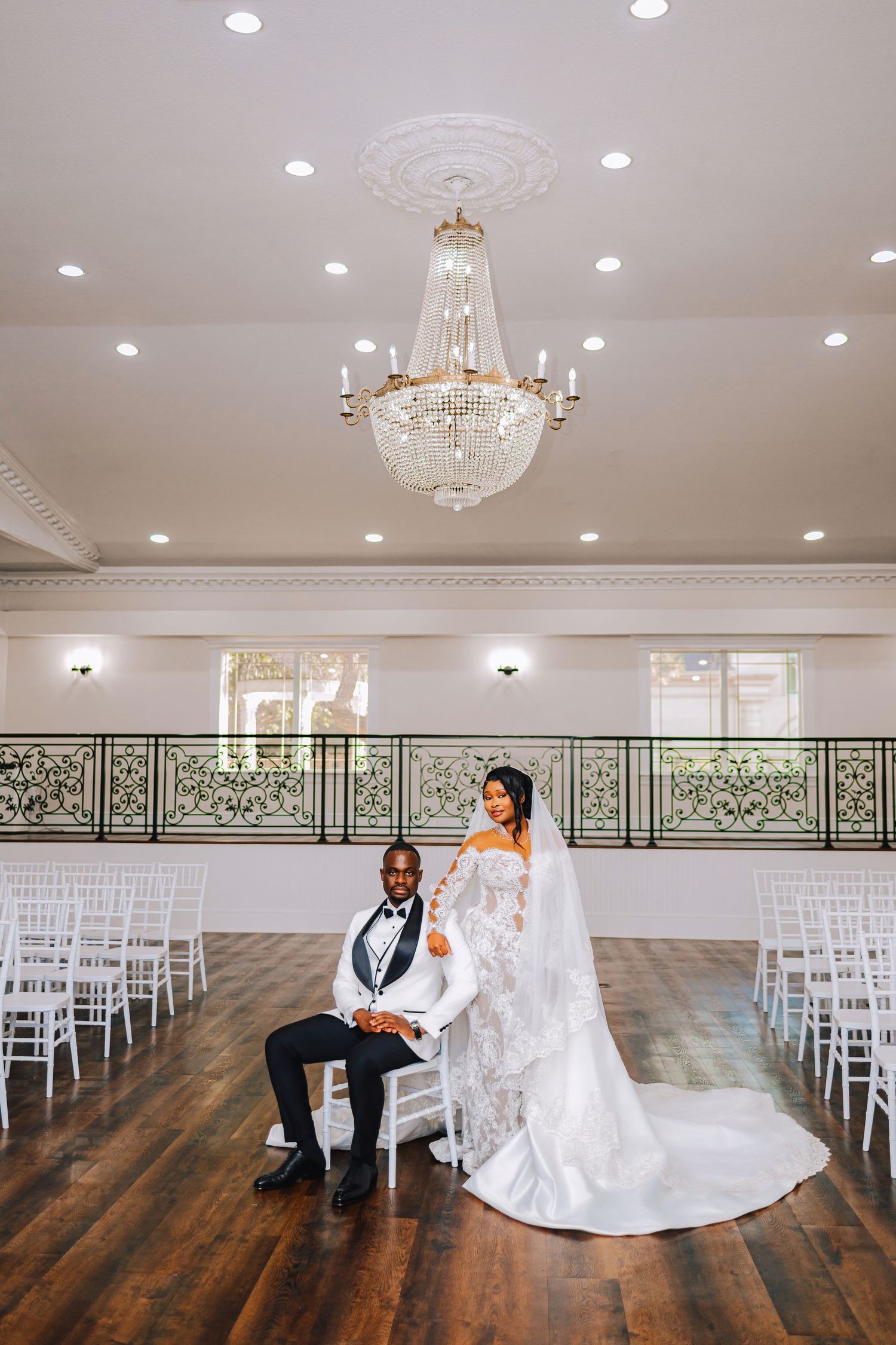Bride in a lace wedding gown with a long veil stands beside a seated groom in a white tuxedo jacket in an elegant ballroom with a chandelier