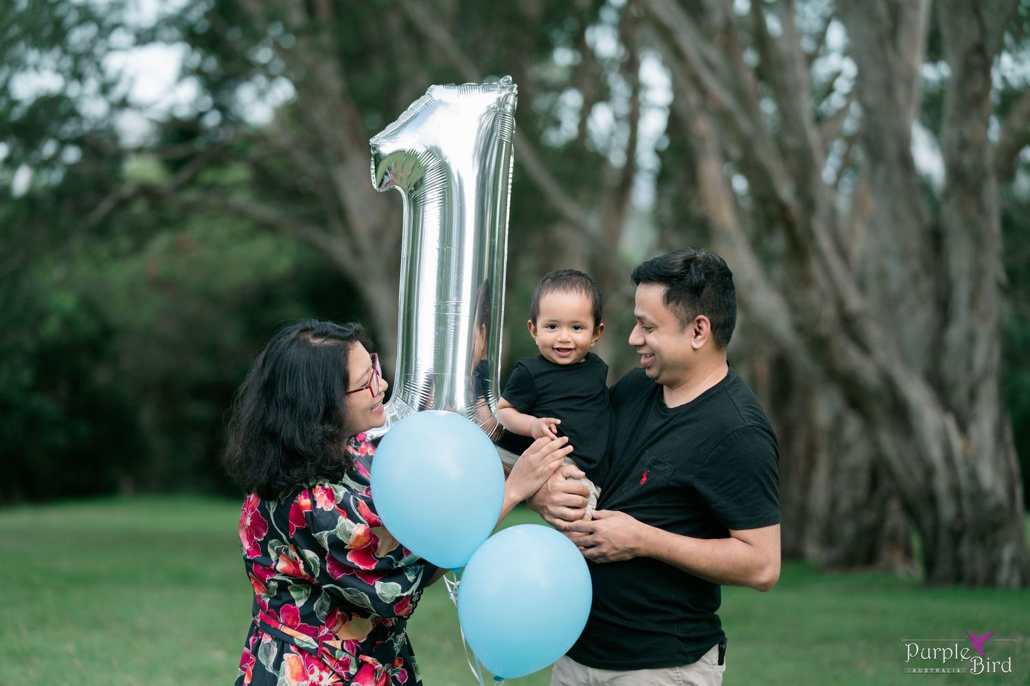 Parents hold their toddler beside blue and number one balloons in the park - first birthday family photoshoot