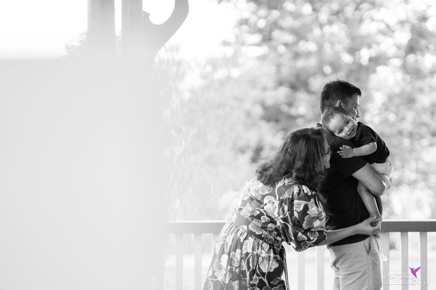 Black and white photo of a mother kissing the father as he holds their child on a porch - timeless family portraits