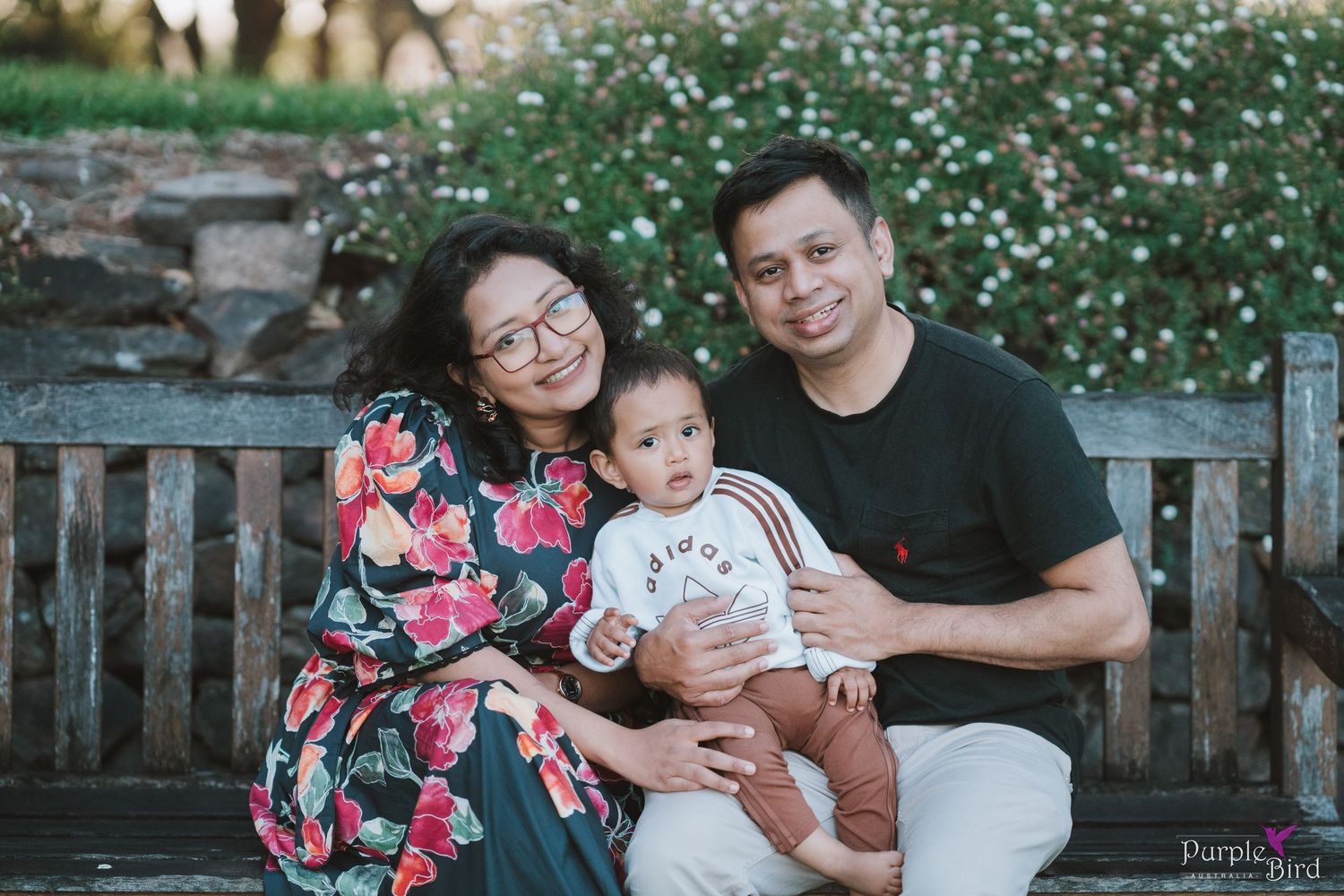 Parents with their baby on a wooden bench beside white flowers - family photoshoot outfit ideas in soft florals