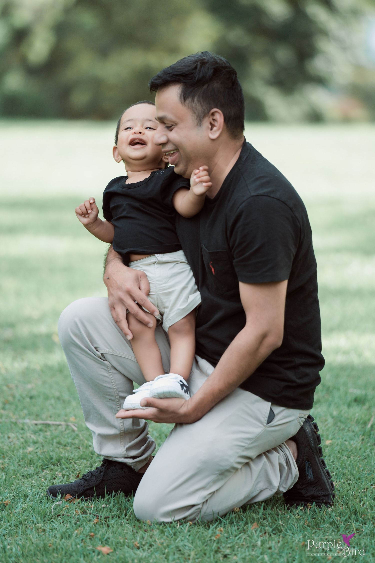 Father kneels on the grass laughing with his toddler son - candid outdoor family photos by a Wollongong family photographer