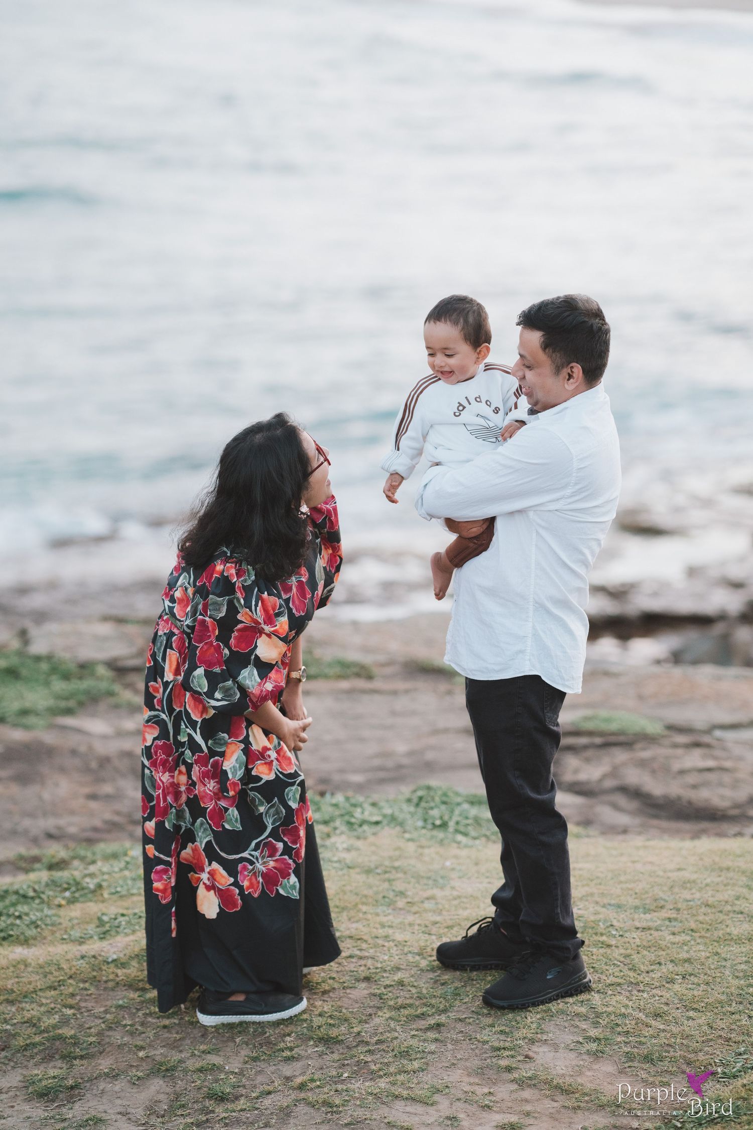 Father holds a smiling toddler facing the mother on a grassy coastal bluff above the ocean - coastal family photoshoot NSW