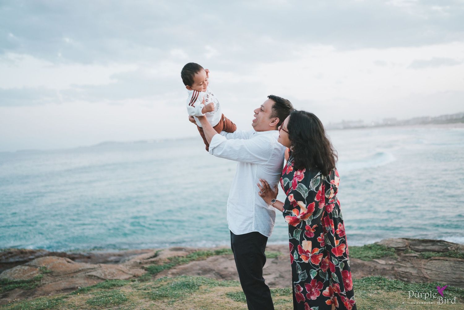 Father lifts the baby beside the mother on a rocky coastline at dusk - golden hour family photography by the ocean