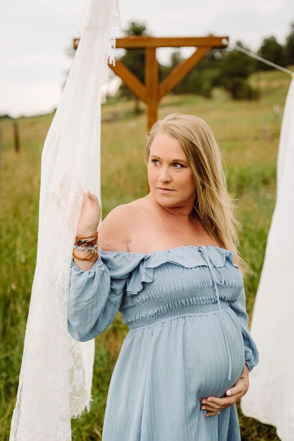 Pregnant woman in blue off-shoulder dress holding white fabric outdoors in a green field maternity photo session.