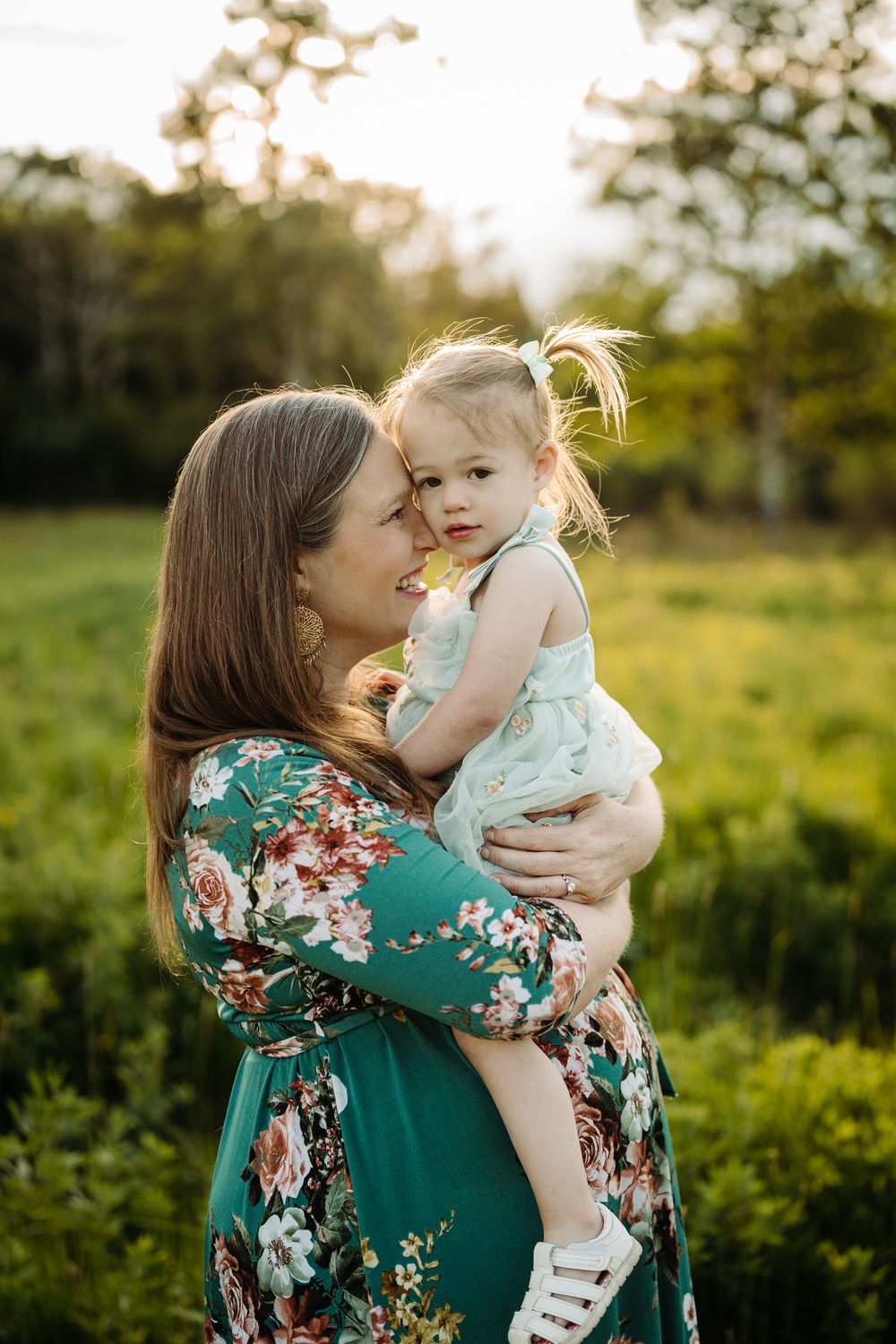 A mother in a floral green dress holds her toddler daughter in a sunny green field at golden hour.
