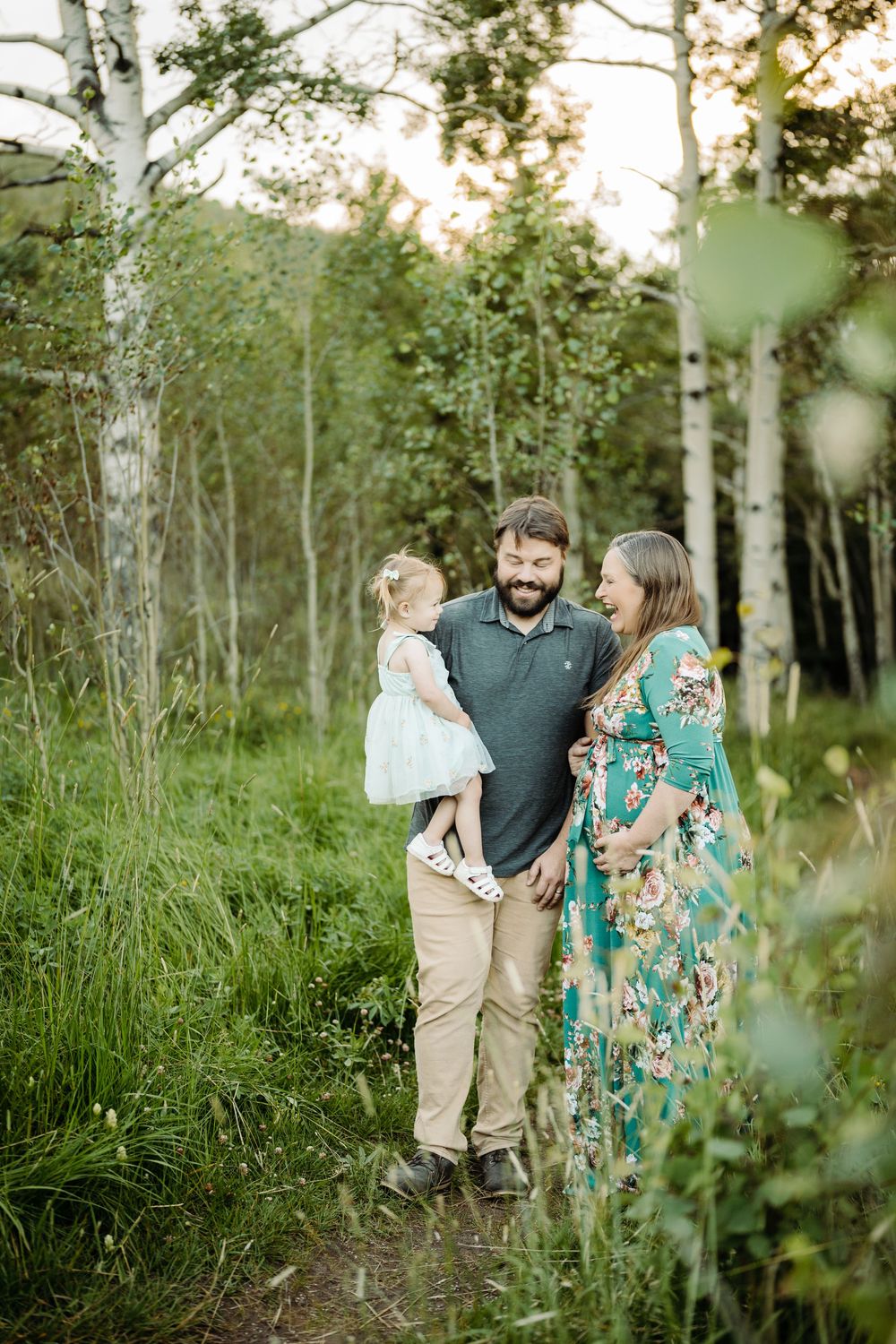 Bearded father holds toddler girl while pregnant mother in floral dress laughs in birch tree forest path.