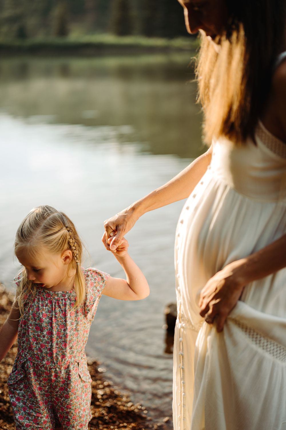 Pregnant woman in white dress holds young blonde girl's hand near a sunlit lake outdoors.