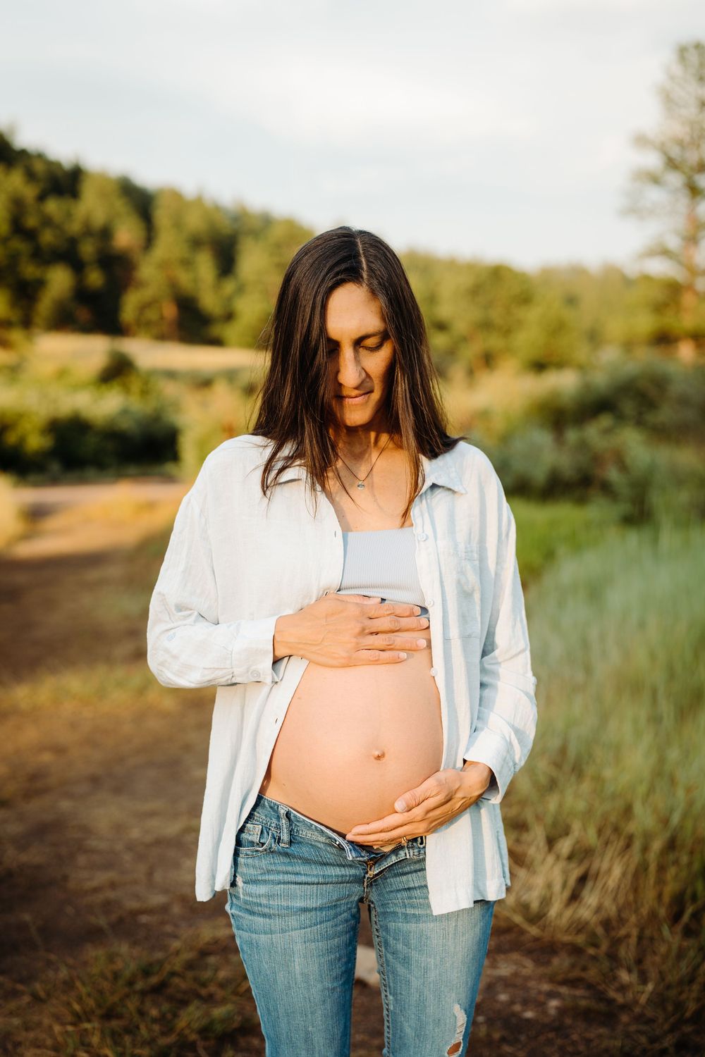 Pregnant woman in white shirt and jeans cradling her baby bump outdoors in a sunny natural setting.