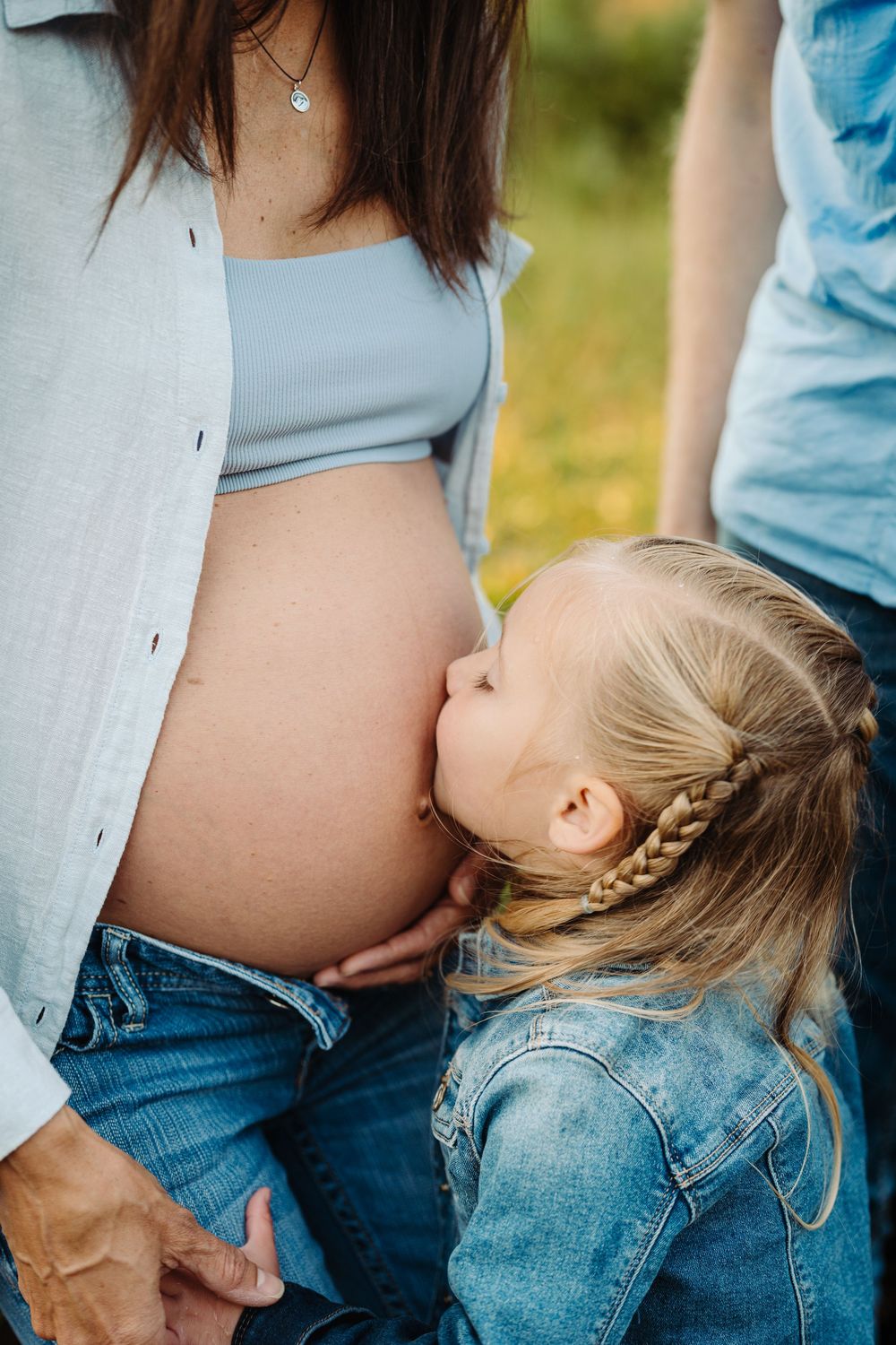 A young girl with a braid kisses her pregnant mothers bare baby bump outdoors.