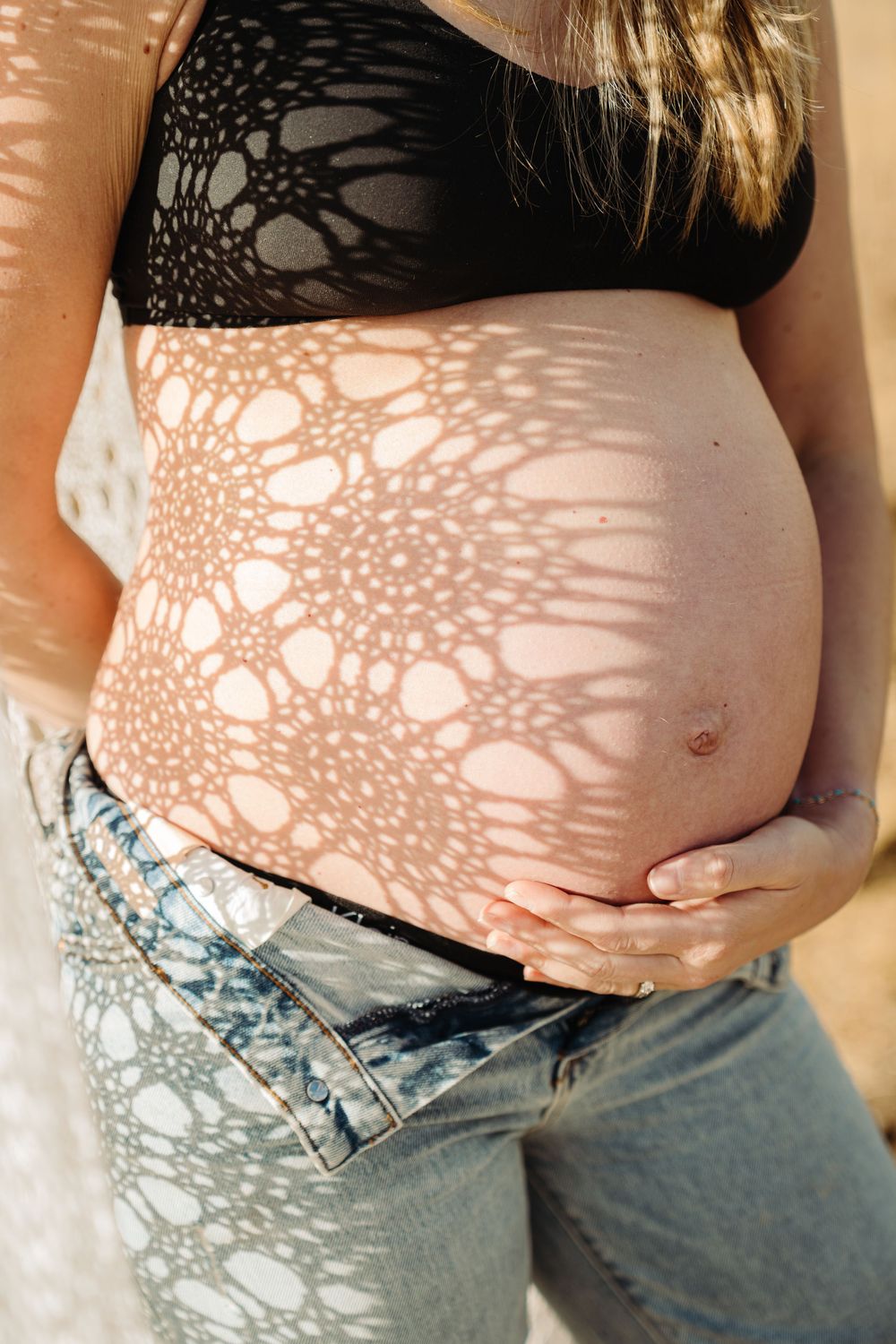 Close-up of pregnant belly in jeans with lace shadow pattern in natural sunlight.