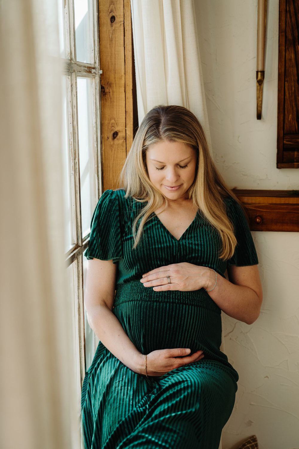 Pregnant woman in green velvet dress cradling baby bump by window.