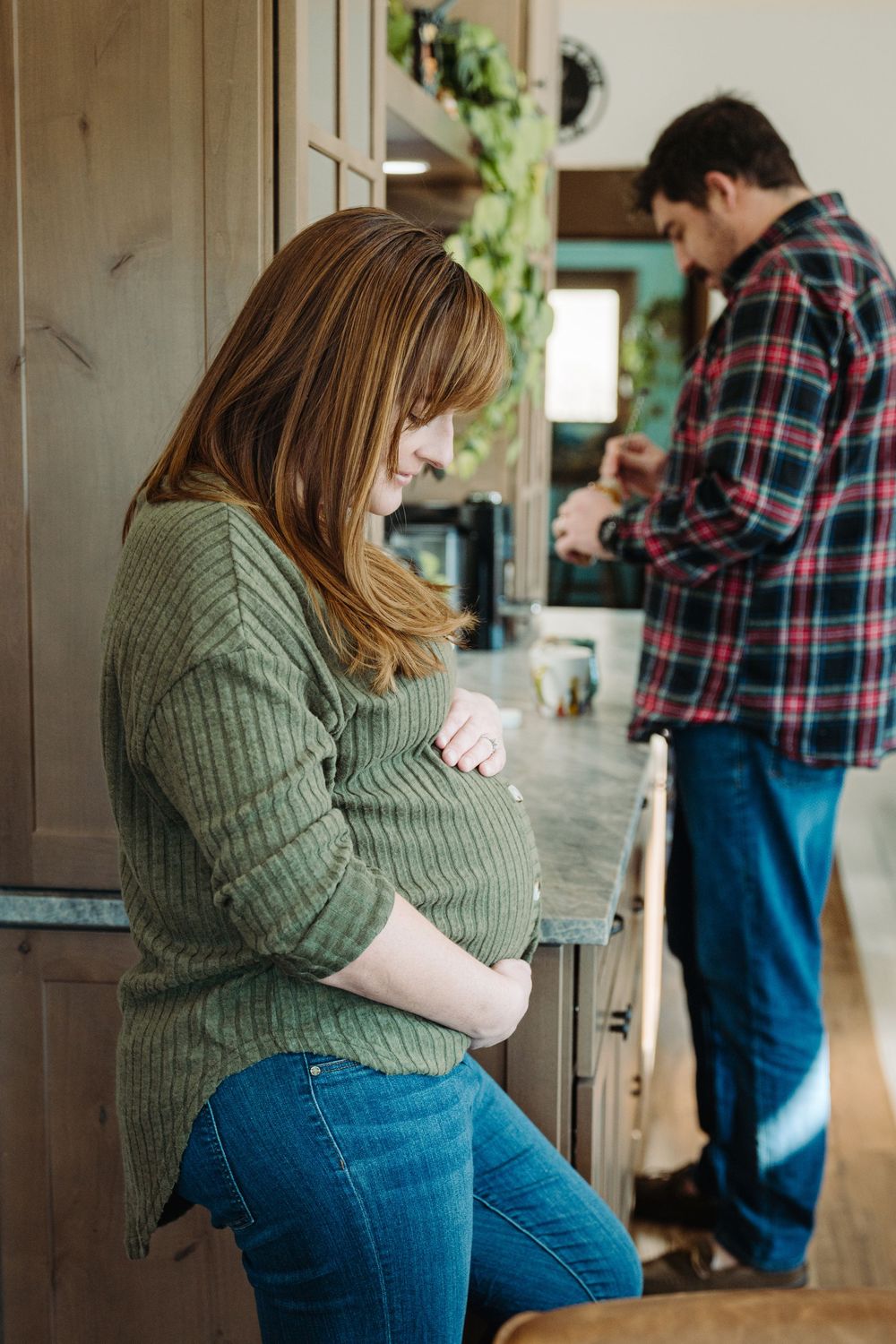 Pregnant woman in green sweater cradling her baby bump in a cozy kitchen while a man works in the background.