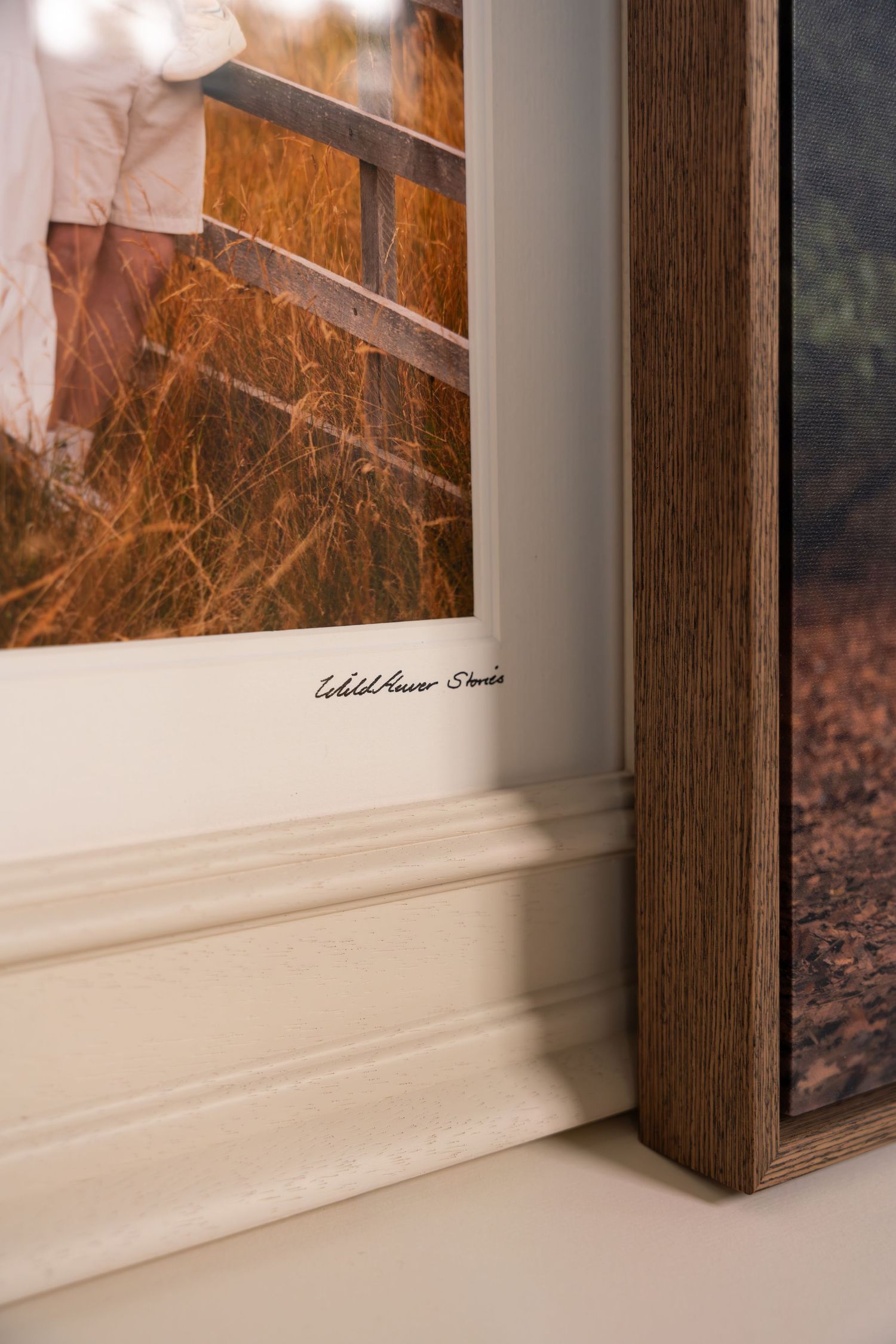Corner of a white-matted framed photograph signed Wildflower Studio, resting on a ledge beside a wooden frame against a cream wall.