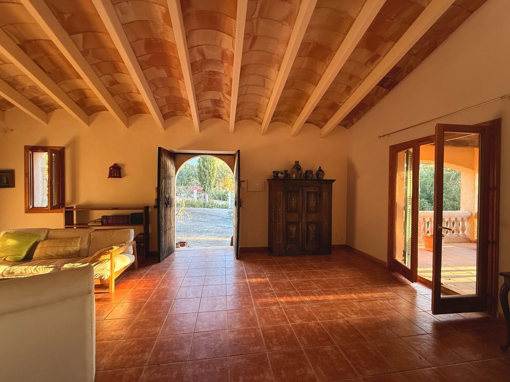 Spacious rustic living room with terracotta tile floors, vaulted wood-beam ceiling, open double doors, and a view of a sunny garden.