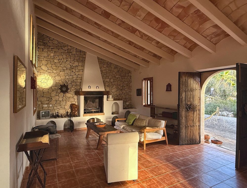 Rustic living room with exposed wooden beams, stone fireplace, terracotta tile floor, cozy seating and an open arched door to the garden.